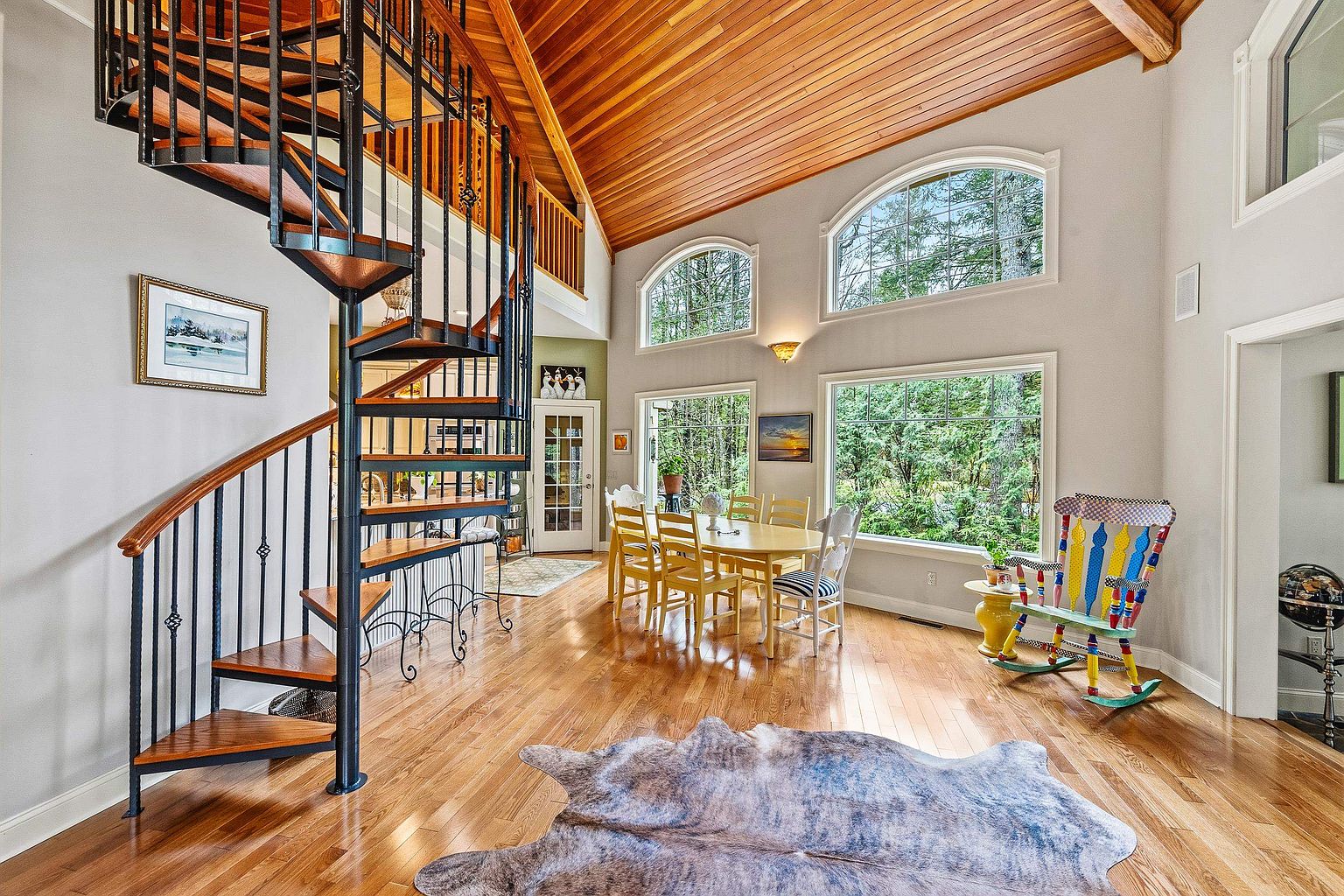 This bright and airy dining area features a striking spiral staircase with wood treads and black metal railings as a central architectural focal point. The room is characterized by high vaulted ceilings with warm wood paneling, large windows that flood the space with natural light, and polished hardwood flooring. A colorful, whimsical rocking chair and a rustic cowhide rug add unique character to this inviting, open-concept living space.