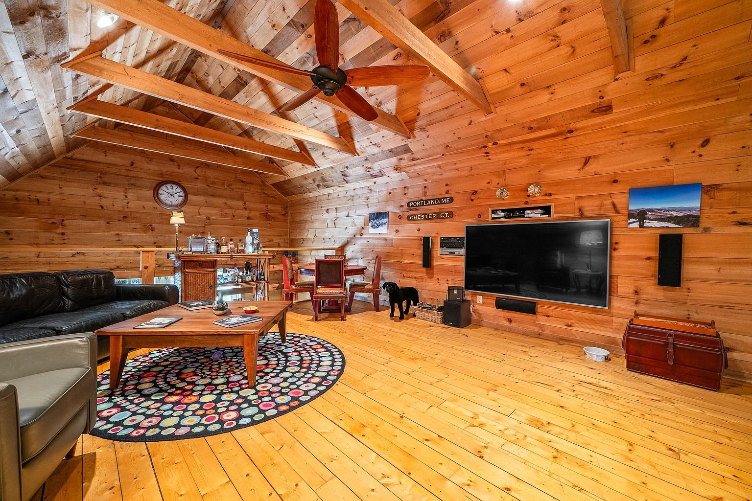 This spacious, rustic-style living room features warm wood-paneled walls and a vaulted ceiling with exposed beams and a large ceiling fan. The room is furnished with a black leather sofa, a wooden coffee table atop a colorful circular rug, and a large wall-mounted television, creating a cozy and inviting entertainment space. A small bar area and a dining table are tucked into the background, while a black dog stands near the television, adding a touch of homey character to the room.