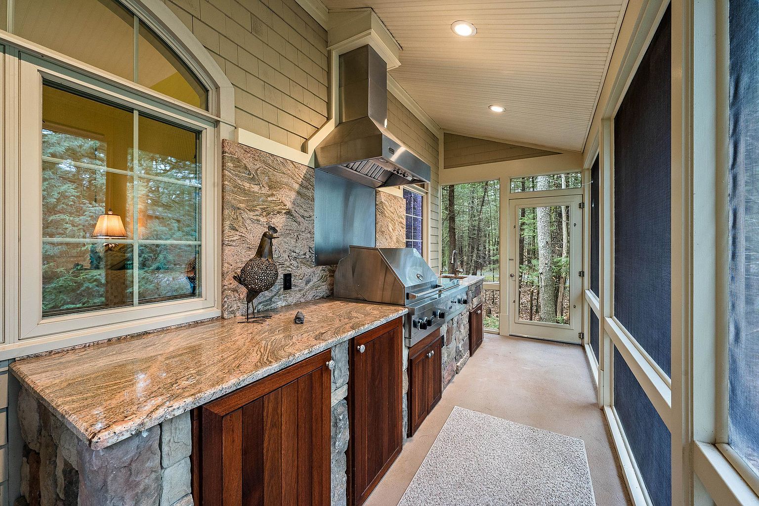 This screened-in porch features a high-end outdoor kitchen complete with a stainless steel grill, range hood, and granite countertops set against a rustic stone base. The space is enclosed by large screens that offer a serene, immersive view of the surrounding forest, creating a perfect blend of indoor comfort and outdoor nature. The warm wood cabinetry and stone accents provide a sophisticated, cozy atmosphere ideal for al fresco dining and entertaining.