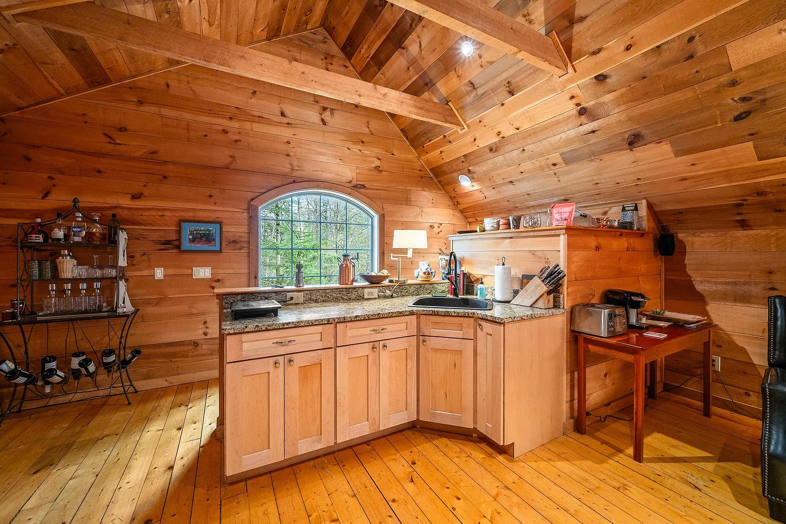 This cozy, rustic kitchen is nestled within an attic-style space featuring warm, natural wood paneling on the walls and vaulted ceiling. The L-shaped layout includes light-toned wooden cabinetry, granite countertops, and a small side table for appliances, all illuminated by a large arched window that brings in natural light. A separate metal wine rack and bar area to the left complete this charming, cabin-inspired culinary nook.