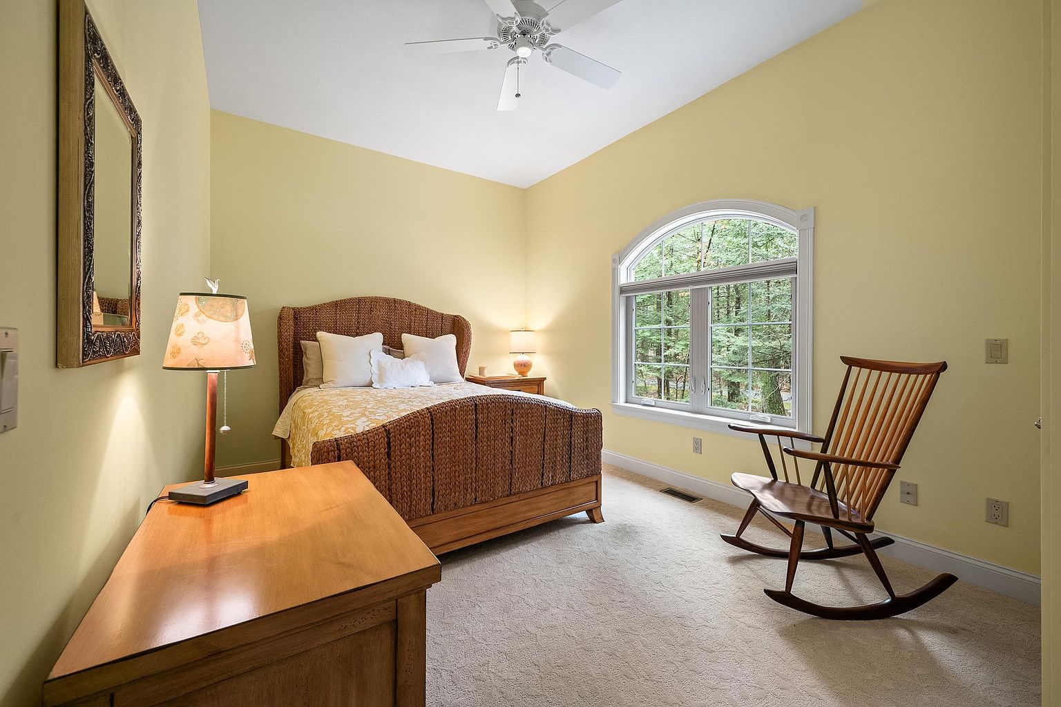 This cozy guest bedroom features a warm, inviting atmosphere with soft yellow walls and plush carpeting. The room is anchored by a woven wicker bed frame and complemented by a classic wooden rocking chair positioned near a large, arched window that brings in natural light. A wooden desk and lamp in the foreground add functionality, creating a comfortable and serene retreat.