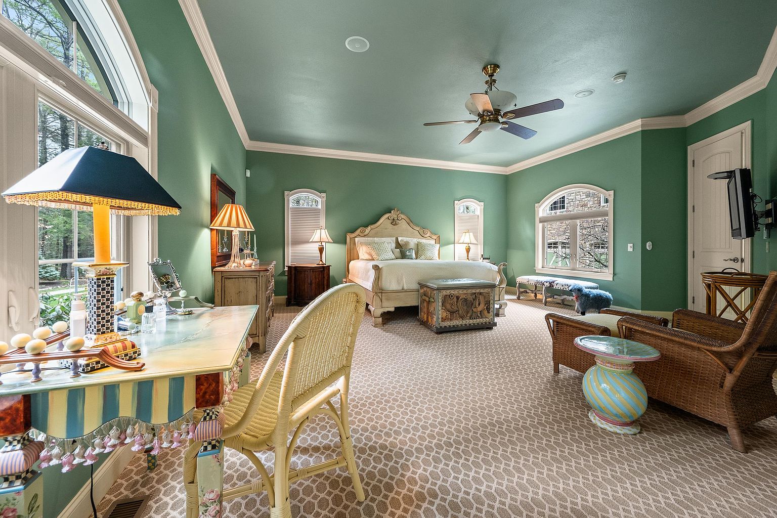 This spacious primary bedroom features a distinctive green color palette on both the walls and ceiling, complemented by a patterned carpet and arched windows. The room is furnished with an ornate bed frame, a whimsical vanity desk, and a cozy seating area with wicker furniture. The overall aesthetic is eclectic and traditional, offering a bright and inviting atmosphere with ample natural light.
