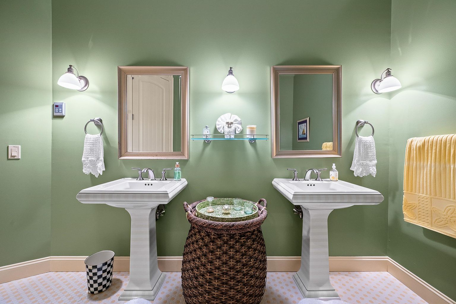 This bright and inviting bathroom features a double pedestal sink vanity set against a soft sage green wall. The space is accented by two framed mirrors, matching wall sconces, and a central woven basket serving as a decorative storage piece. The room is finished with a patterned tile floor and a yellow towel, creating a clean and cheerful aesthetic.