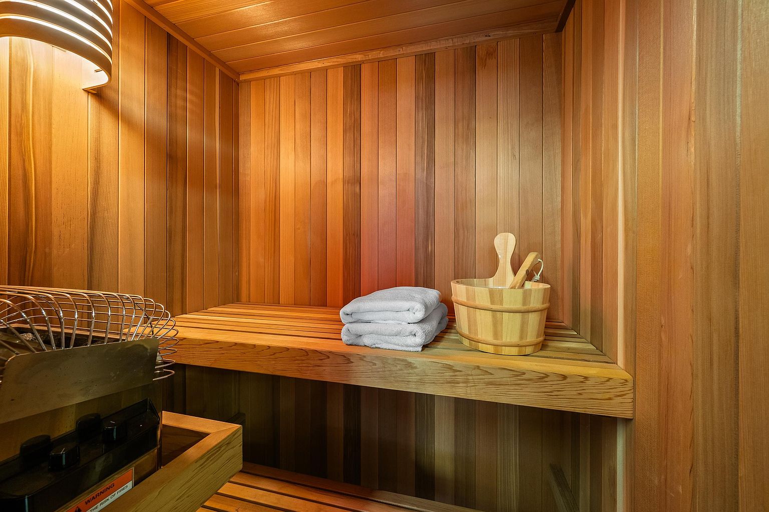 This image showcases a compact, wood-paneled sauna featuring warm cedar walls and a built-in bench. A wooden bucket with a ladle sits on the bench next to neatly folded white towels, while a metal heater is visible in the foreground. The warm lighting and natural wood textures create a cozy, spa-like atmosphere perfect for relaxation.