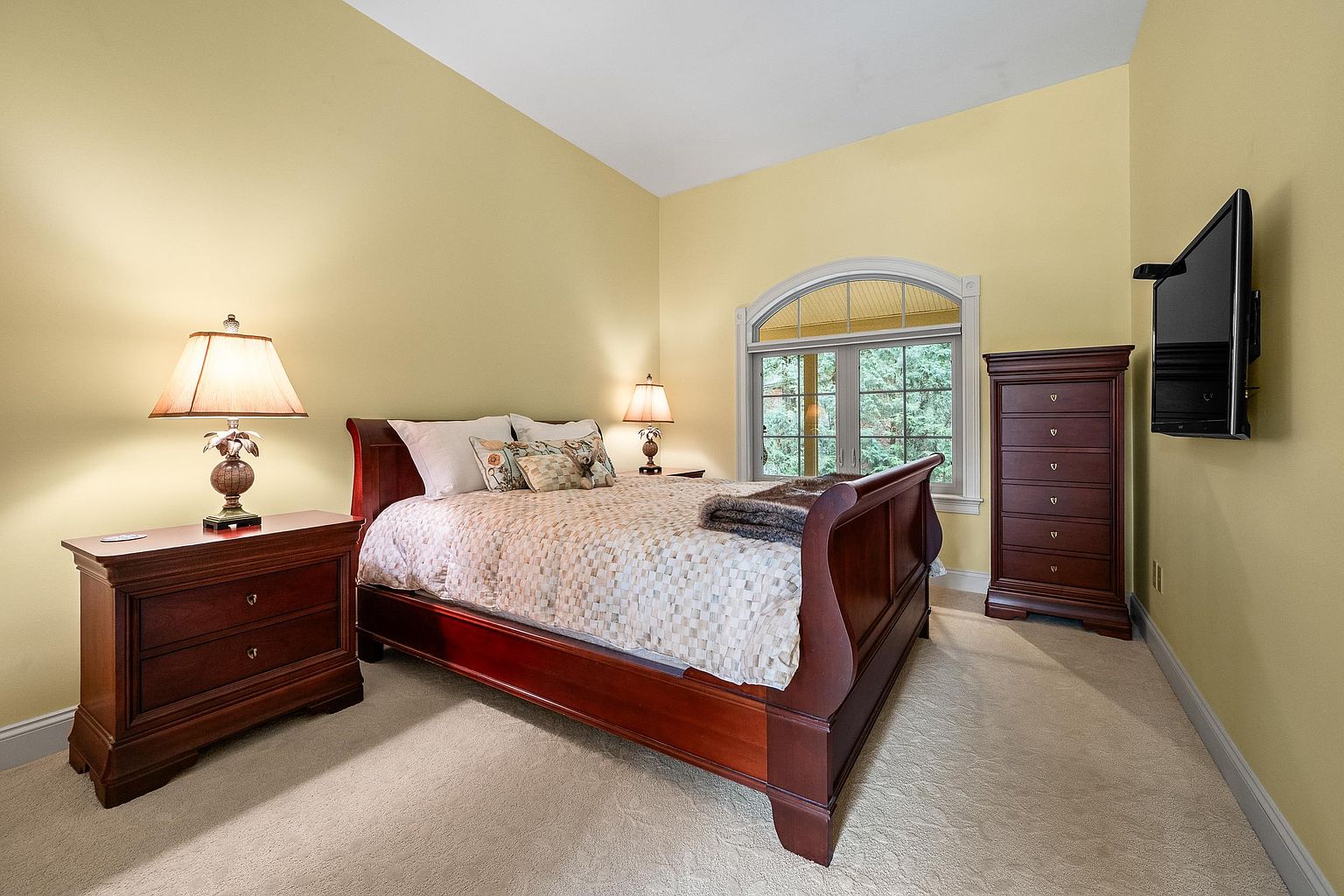 This cozy bedroom features a rich, dark wood sleigh bed as the centerpiece, complemented by matching nightstands and a tall dresser. The room is painted in a warm, inviting yellow tone and includes a large arched window that allows natural light to filter in. A wall-mounted television and soft, neutral carpeting complete this comfortable and traditional living space.