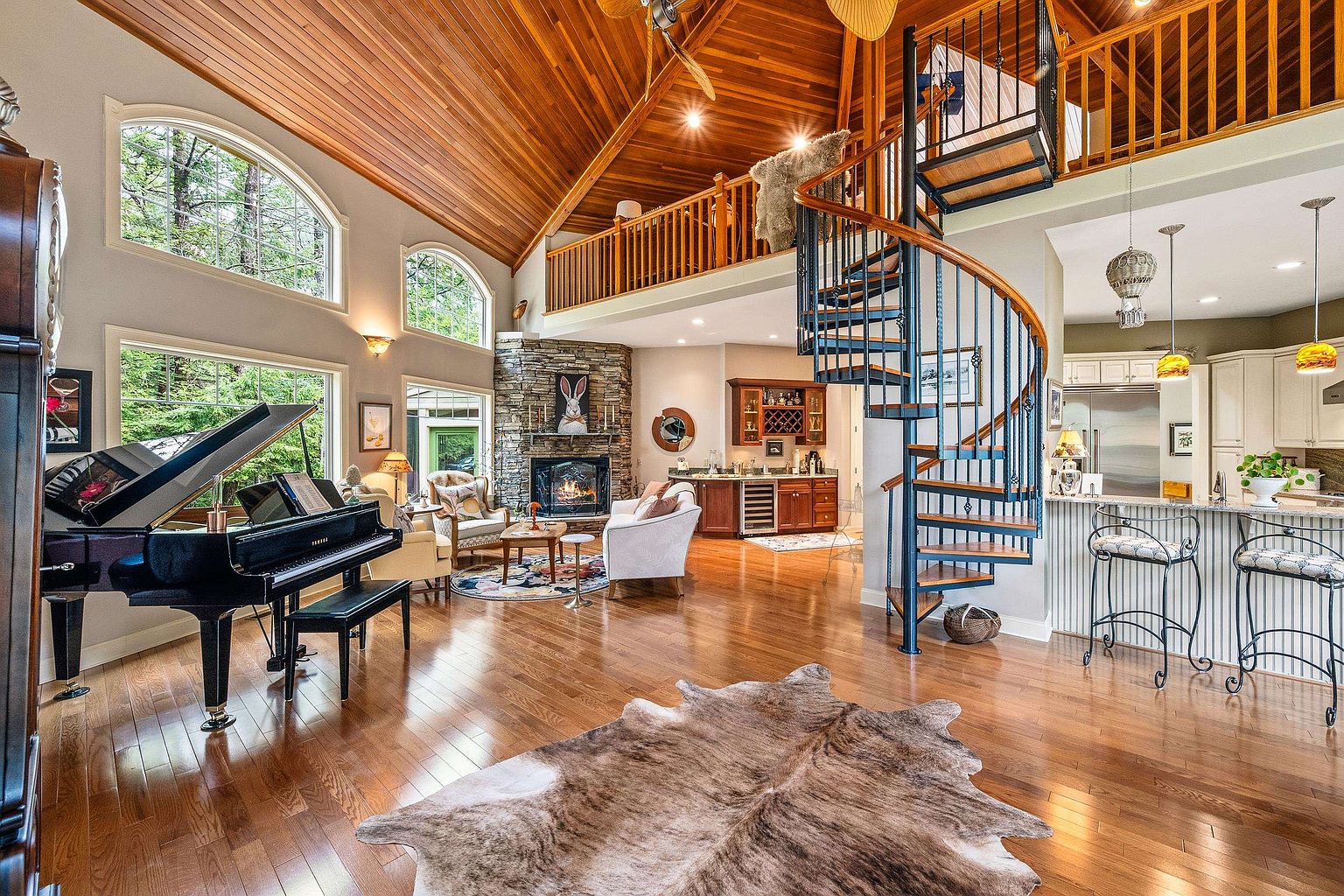 This expansive, open-concept living area features a dramatic vaulted wood-paneled ceiling and a striking spiral staircase leading to a loft. The space is anchored by a stone fireplace, a grand piano, and warm hardwood flooring, creating a sophisticated yet cozy atmosphere. Large arched windows flood the room with natural light, highlighting the seamless transition between the living space, a wet bar, and the adjacent kitchen.
