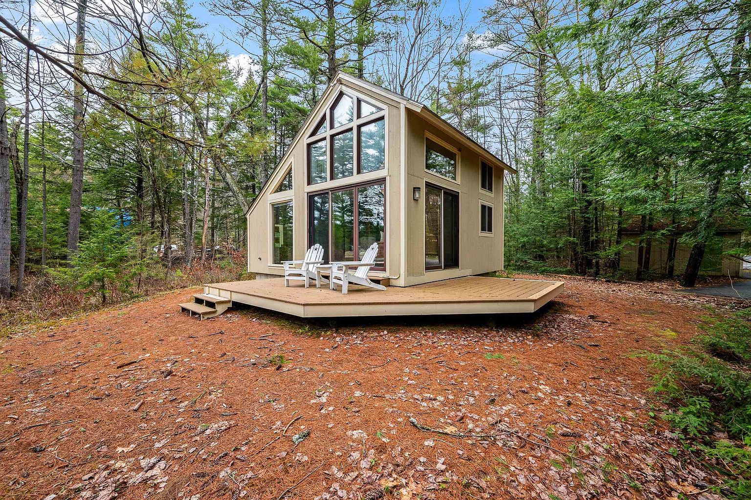 This charming, modern A-frame cabin is nestled in a serene, wooded setting, featuring a large, elevated wooden deck with two white Adirondack chairs. The structure boasts expansive floor-to-ceiling windows that flood the interior with natural light, reflecting the surrounding forest. The exterior is finished in a neutral, earthy tone, creating a peaceful and inviting retreat atmosphere.