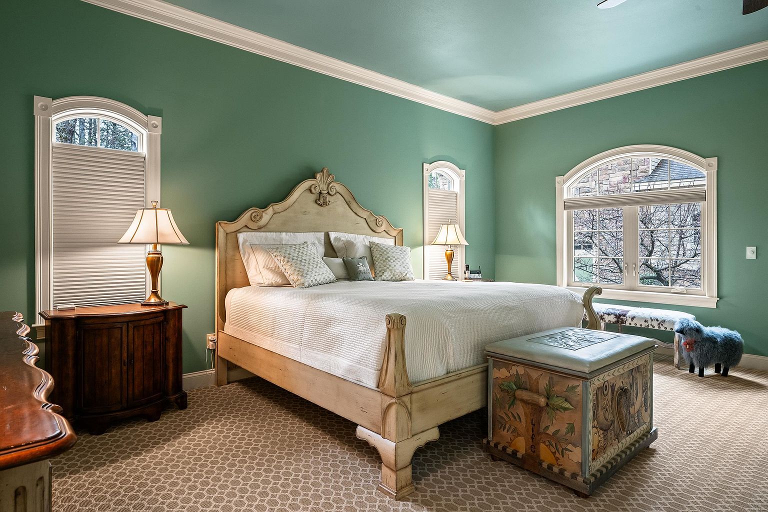 This elegant primary bedroom features a light-toned, ornate wooden bed frame as the centerpiece, complemented by matching nightstands and a decorative painted storage chest at the foot of the bed. The room is painted in a soothing sage green with crisp white crown molding and arched windows that allow for natural light. A patterned carpet adds texture to the space, while a whimsical blue sheep ottoman provides a playful touch to the sophisticated decor.