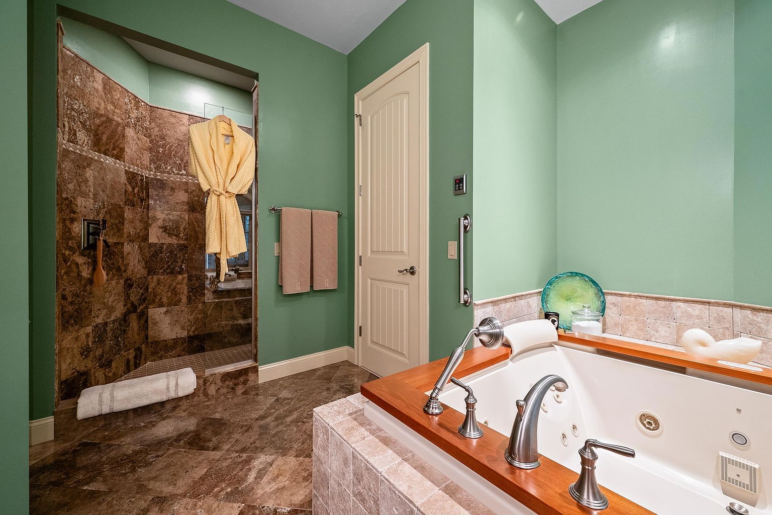 This primary bathroom features a luxurious jetted soaking tub with a rich wood trim surround and a separate walk-in shower finished in dark, textured stone tiles. The walls are painted a soft sage green, creating a calming atmosphere, while the floor is laid with matching stone tile. A yellow bathrobe hangs in the shower, adding a touch of warmth to the space, which is accented by polished chrome fixtures and a grab bar for accessibility.