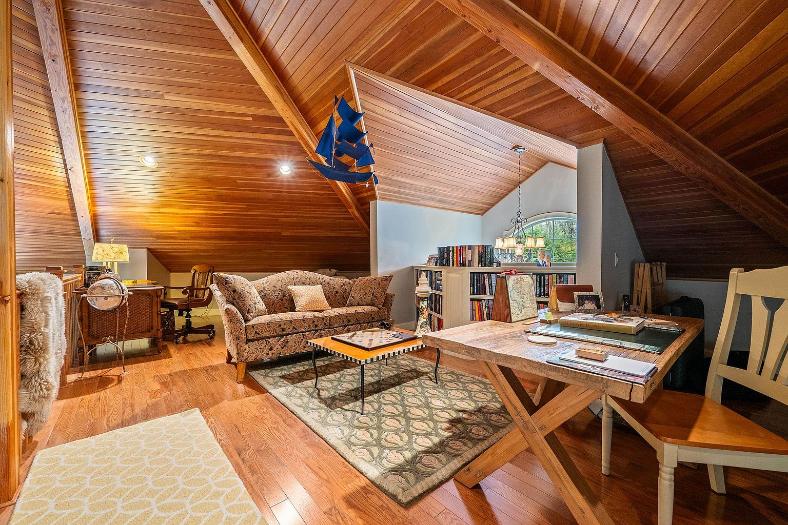This cozy, wood-paneled attic space serves as a multi-functional study and lounge area, featuring vaulted ceilings and warm, natural wood tones. The room is furnished with a patterned sofa, a central coffee table, and a sturdy wooden desk, creating a comfortable and inviting atmosphere. A large bookshelf and a decorative model ship add character to the space, while the warm lighting enhances the rustic, cabin-like aesthetic.