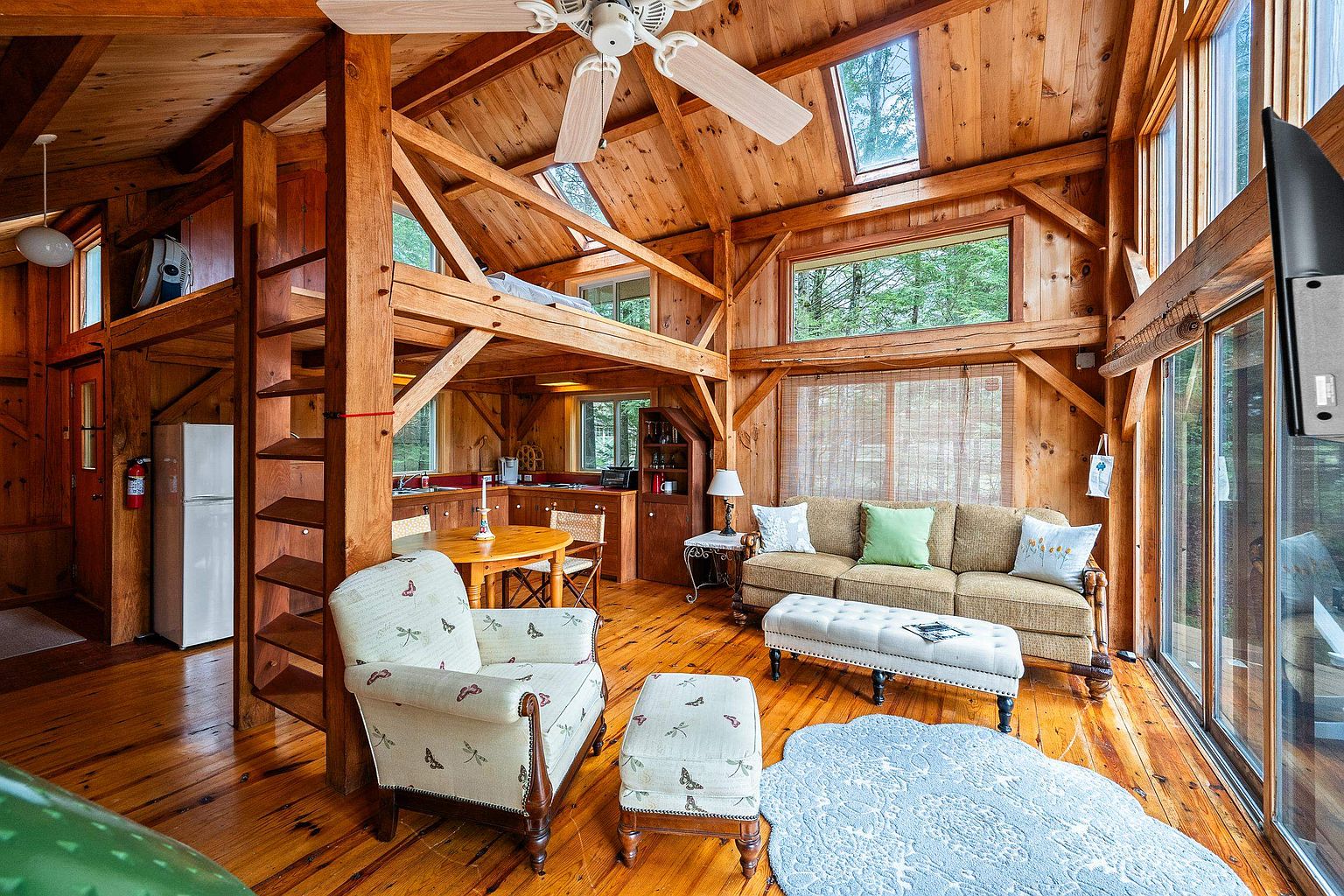 This rustic, open-concept living space features warm wood-paneled walls, exposed timber beams, and a vaulted ceiling with skylights that flood the room with natural light. The area includes a cozy seating arrangement with a patterned armchair, a tufted ottoman, and a sofa, all set upon a light blue area rug. A wooden dining table and a glimpse of a kitchen area are visible in the background, emphasizing the home's inviting, cabin-like atmosphere.