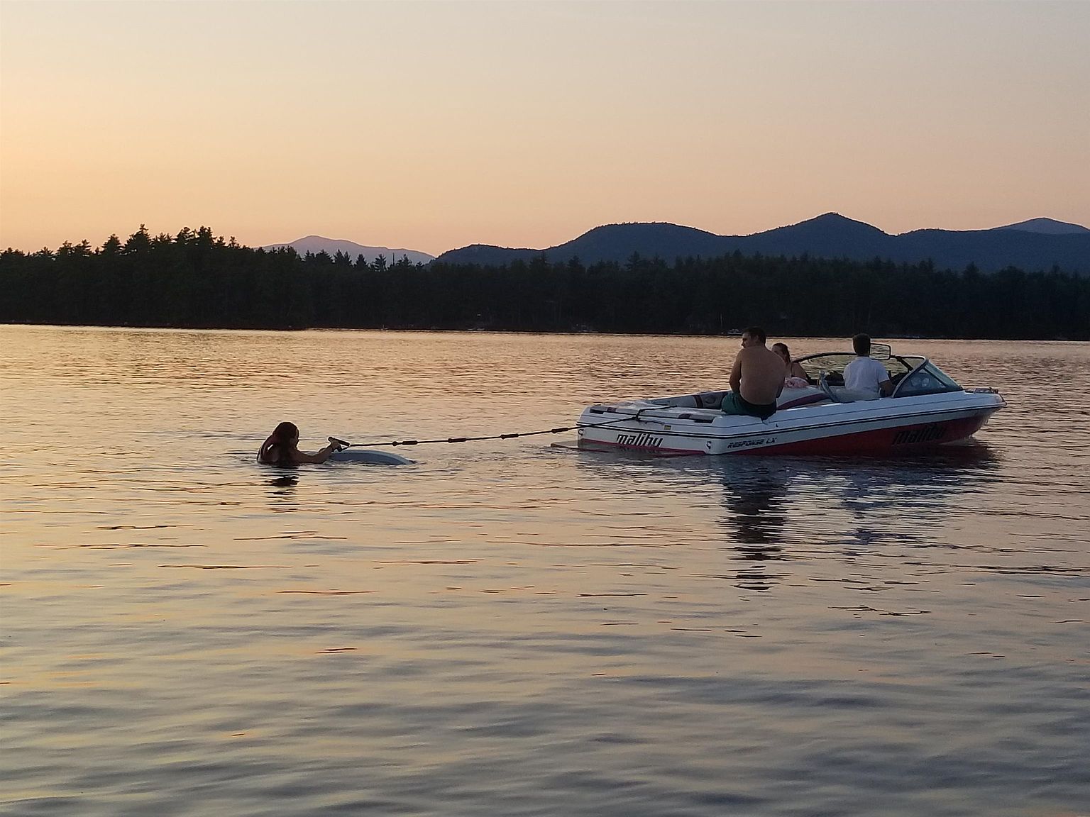 This scenic shot captures a recreational boating scene on a calm lake at sunset, featuring a Malibu motorboat towing a person in the water. The background showcases a dense treeline and rolling mountains, creating a peaceful and idyllic atmosphere perfect for highlighting a waterfront lifestyle. The perspective is a wide, eye-level shot that emphasizes the vastness of the water and the serene evening light.
