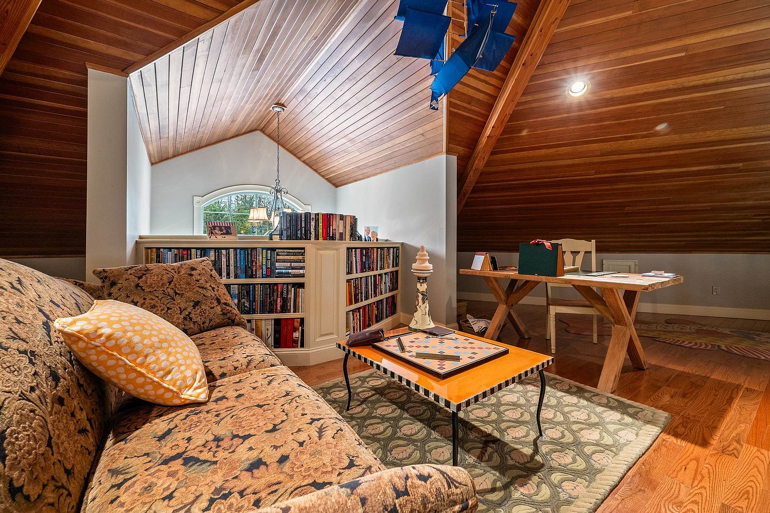 This cozy, wood-paneled attic space serves as a multi-functional reading nook and home office. It features a patterned sofa with a yellow accent pillow, a built-in bookshelf filled with literature, and a rustic wooden desk, all set under a dramatic vaulted ceiling. The warm wood tones and soft lighting create an inviting, scholarly atmosphere perfect for quiet study or relaxation.