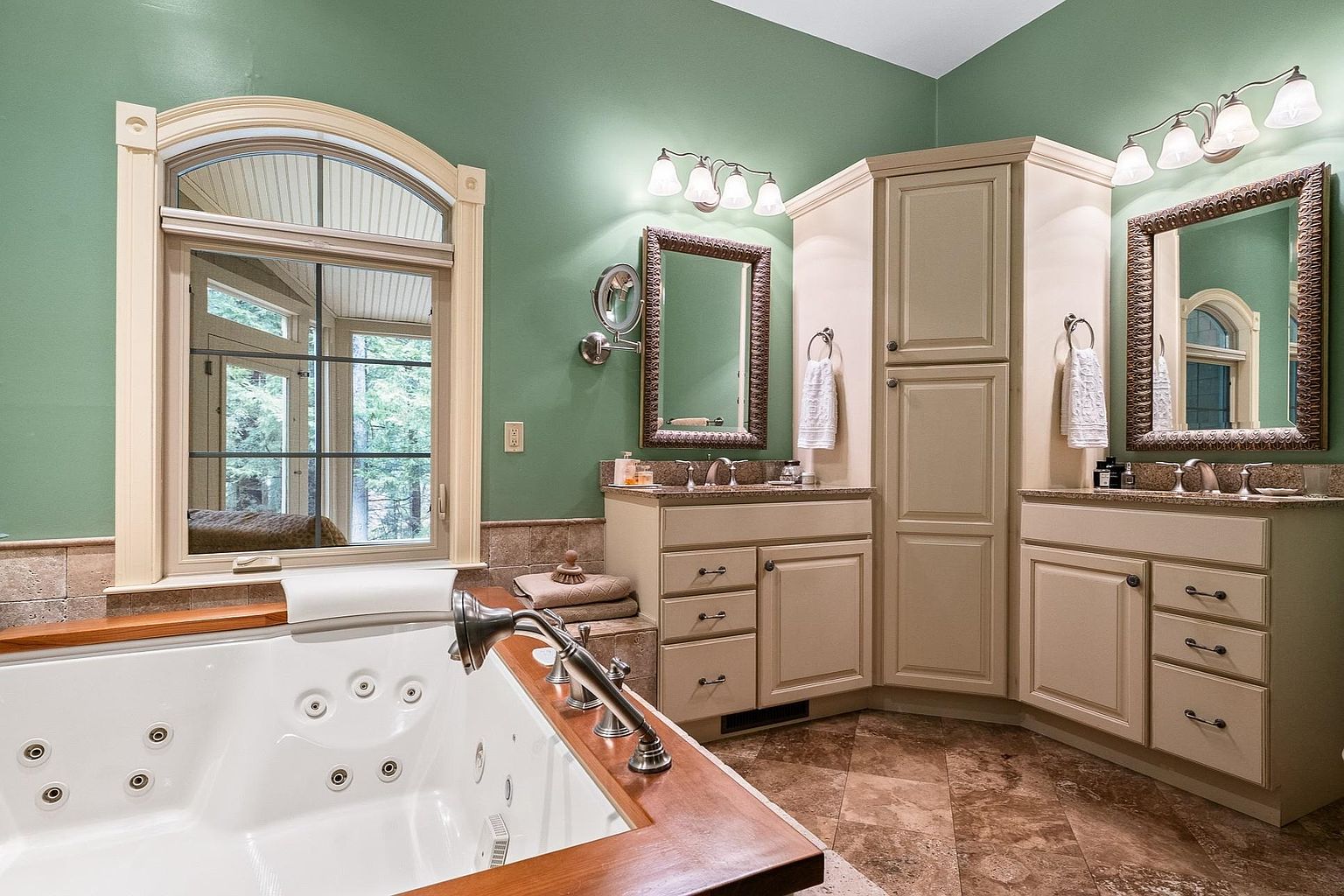 This luxurious primary bathroom features a large jetted soaking tub with a warm wood surround, positioned next to a grand arched window that brings in natural light. The space includes a dual-vanity setup with cream-colored cabinetry, granite countertops, and ornate framed mirrors, all set against a soothing sage green wall color. The combination of tile flooring and elegant lighting fixtures creates a serene, spa-like atmosphere perfect for relaxation.
