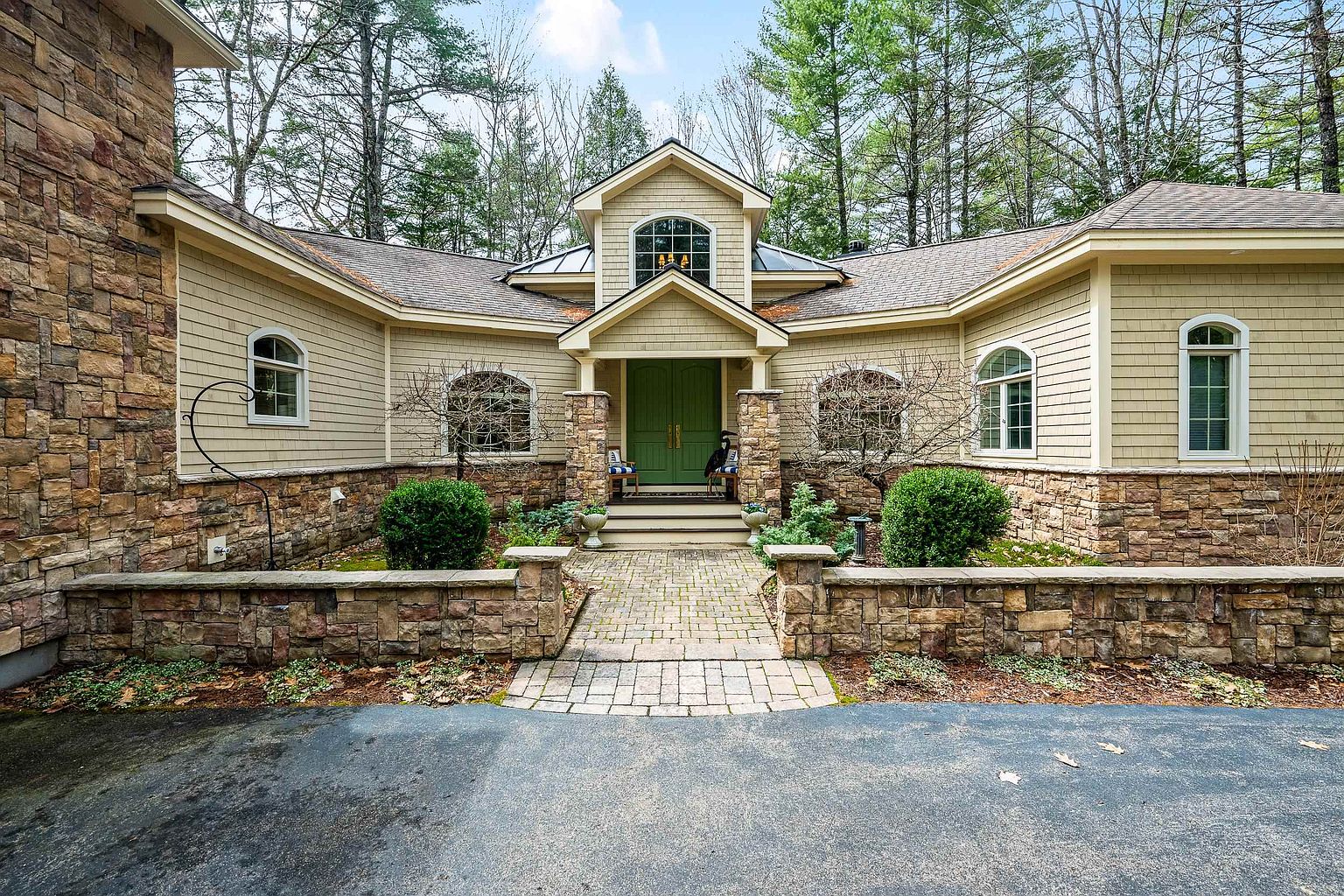 This inviting front exterior features a symmetrical design with a prominent stone facade, light-colored horizontal siding, and a striking green double-door entryway. A stone-paved walkway leads through a low stone retaining wall to the front porch, framed by manicured shrubs and mature trees. The architectural style blends rustic stone elements with classic suburban charm, creating a warm and welcoming curb appeal.