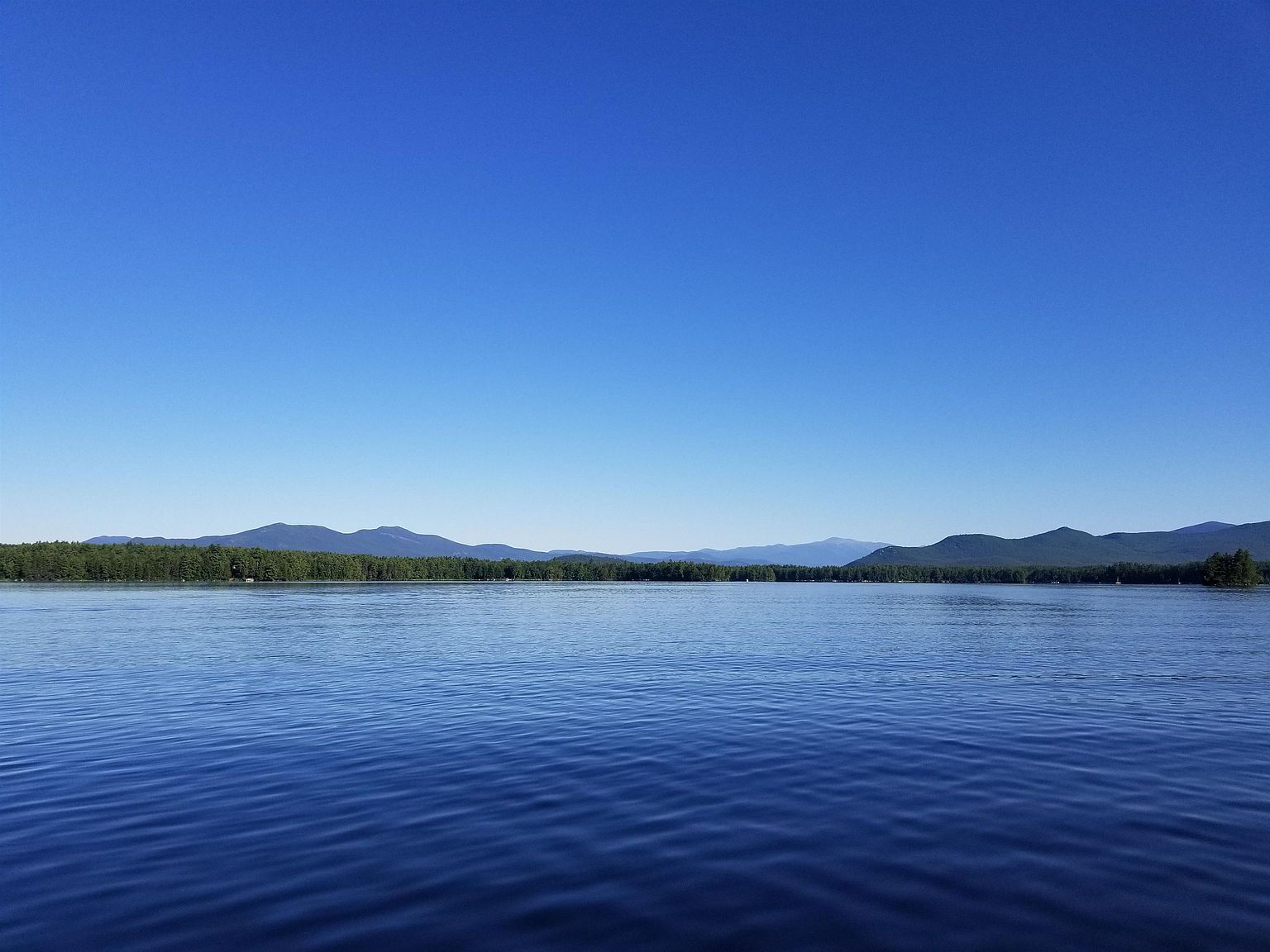 This image captures a serene, wide-angle view of a calm lake surrounded by a dense forest and distant mountain ranges under a clear, bright blue sky. The perspective is from the water's surface, emphasizing the vastness and tranquility of the natural landscape. This scenic view highlights the peaceful waterfront setting, which is a highly desirable feature for potential real estate buyers seeking privacy and natural beauty.