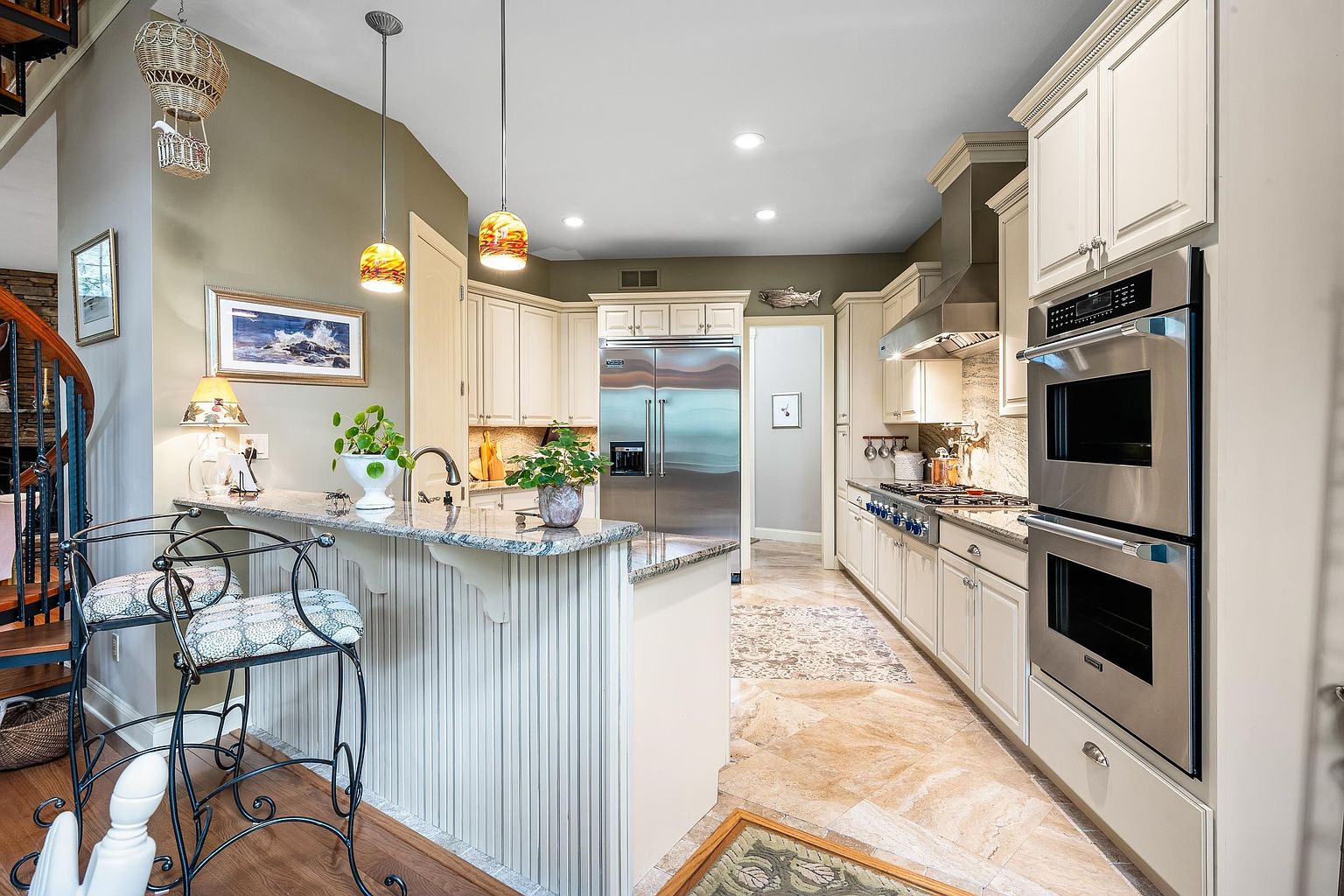 This elegant kitchen features a classic design with cream-colored cabinetry, granite countertops, and high-end stainless steel appliances, including a double wall oven and a large refrigerator. A breakfast bar with two stools provides a casual dining space, while warm pendant lighting and a neutral color palette create an inviting atmosphere. The perspective is a wide-angle shot from the dining area, showcasing the seamless flow into the kitchen and the adjacent hallway.