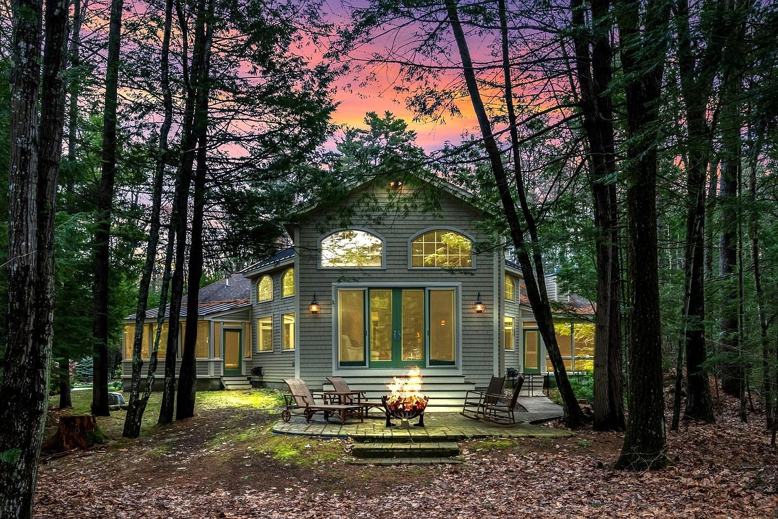 This charming, shingled-siding home is nestled within a dense, tranquil forest, captured at dusk under a vibrant, colorful sunset sky. The rear of the house features a stone patio with a glowing fire pit, surrounded by comfortable outdoor seating, creating an inviting and serene atmosphere. Large, arched windows and glass doors provide a seamless connection between the cozy interior and the natural woodland surroundings.