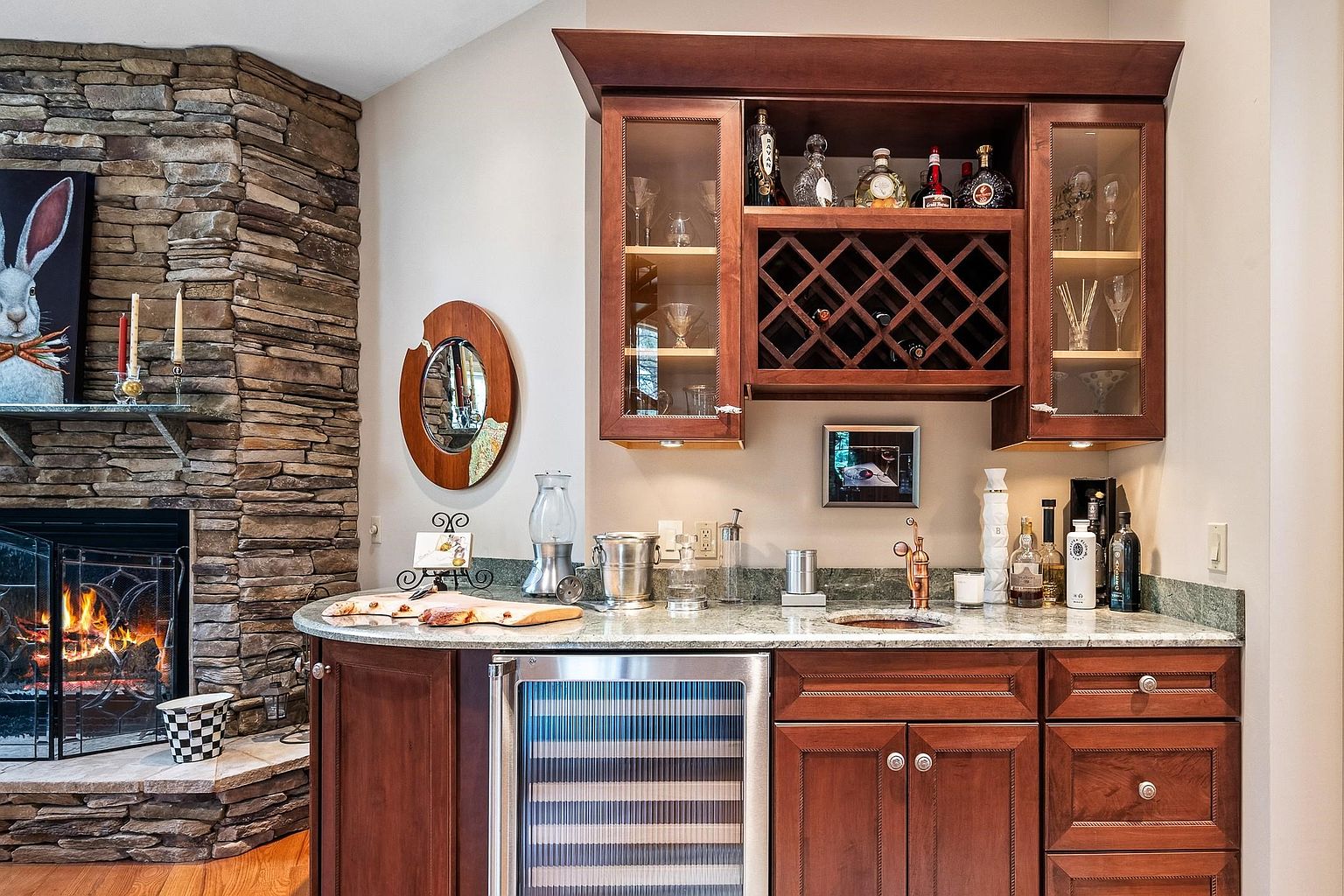 This elegant wet bar area features rich cherry-toned cabinetry, a granite countertop, and a built-in wine refrigerator, seamlessly integrated next to a rustic stone fireplace. The space is accented by glass-front upper cabinets with wine storage, a small sink, and decorative lighting, creating a sophisticated atmosphere for entertaining. The warm wood tones and natural stone textures provide a cozy, high-end aesthetic perfect for a home lounge or living area.