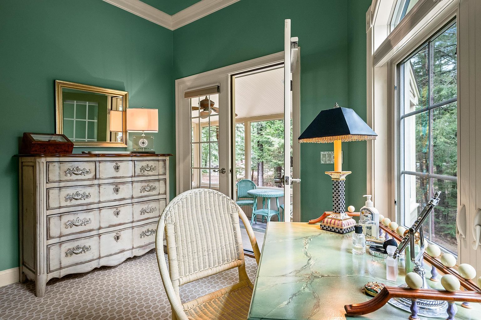 This charming home office or vanity space features vibrant green walls, a distressed white wooden dresser, and a unique painted desk positioned by a large window. A wicker chair adds a touch of coastal elegance, while the open French doors reveal a serene, screened-in porch overlooking a wooded area. The room exudes a tranquil and sophisticated atmosphere, perfect for a quiet workspace or personal retreat.