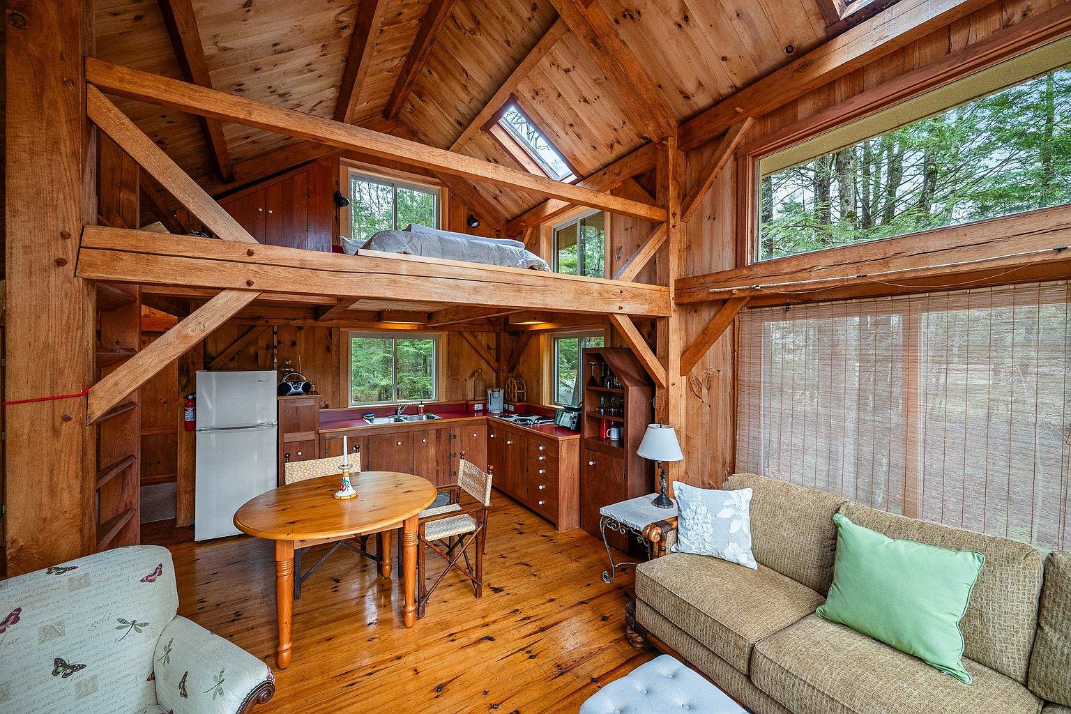 This cozy, rustic cabin interior features an open-concept layout with warm wood paneling, exposed timber beams, and a lofted sleeping area. The space includes a functional kitchen, a dining nook, and a comfortable living area, all bathed in natural light from large windows and a skylight. The overall atmosphere is inviting and serene, perfectly blending traditional craftsmanship with modern comfort.