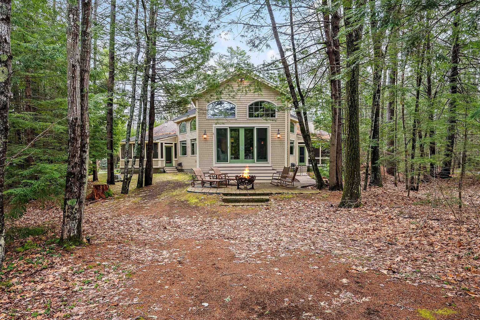 This image captures the rear exterior of a charming, light-colored wood-sided home nestled within a dense, mature forest. The property features a stone patio with a cozy fire pit, surrounded by comfortable outdoor seating, creating an inviting space for relaxation. The architecture includes large windows and a screened-in porch, emphasizing a seamless connection between the home and its tranquil, wooded surroundings.