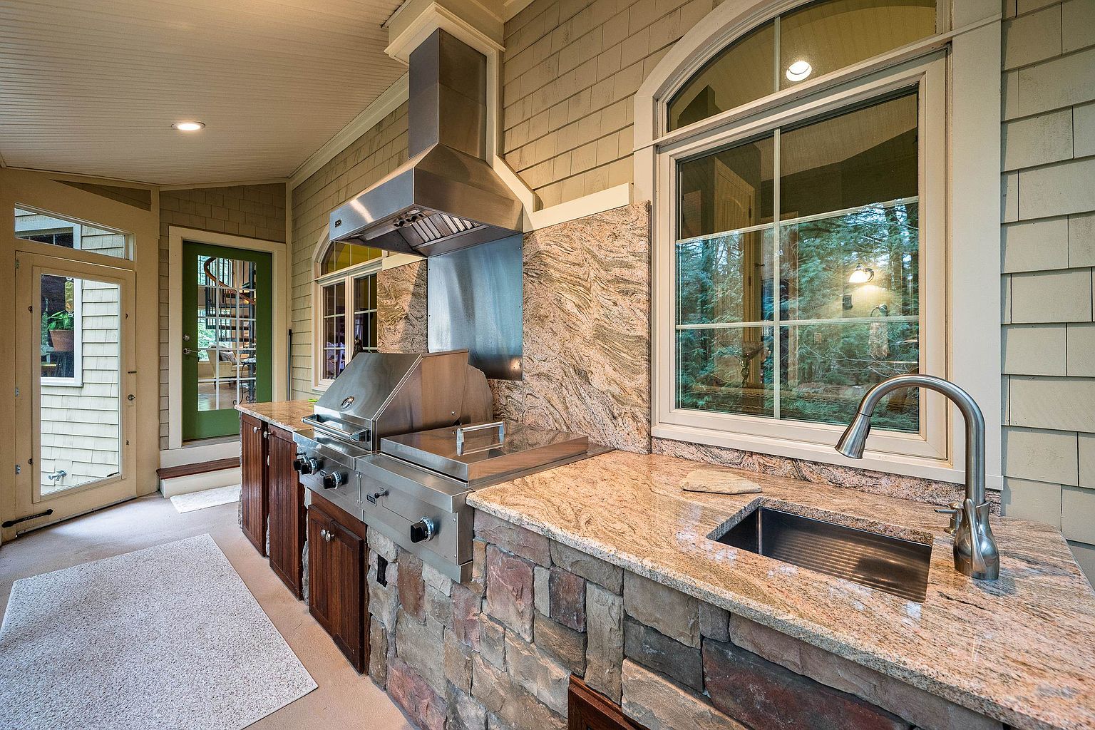 This covered outdoor kitchen area features a high-end stainless steel grill, a stone-clad base, and elegant granite countertops with an integrated sink. The space is framed by light-colored shingle siding and large windows, creating a seamless transition between indoor and outdoor living. The perspective is a wide-angle shot that captures the functional layout and the inviting, rustic-luxury aesthetic of the patio.