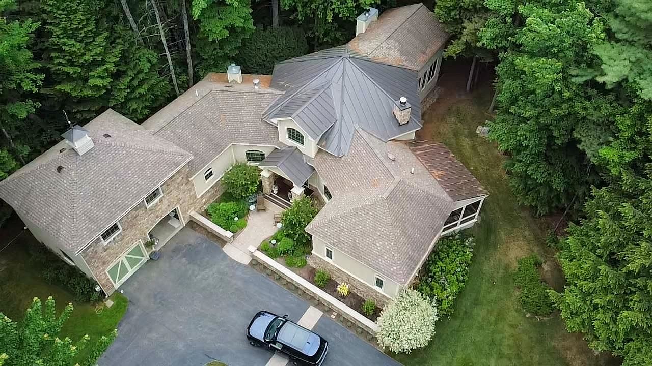 This high-angle aerial view captures a sprawling, multi-gabled luxury residence nestled within a lush, wooded landscape. The home features a blend of stone and stucco siding, a dark metal roof, and a paved driveway with a parked vehicle, all surrounded by mature trees and manicured landscaping. The perspective emphasizes the property's secluded, estate-like setting and its complex architectural design.