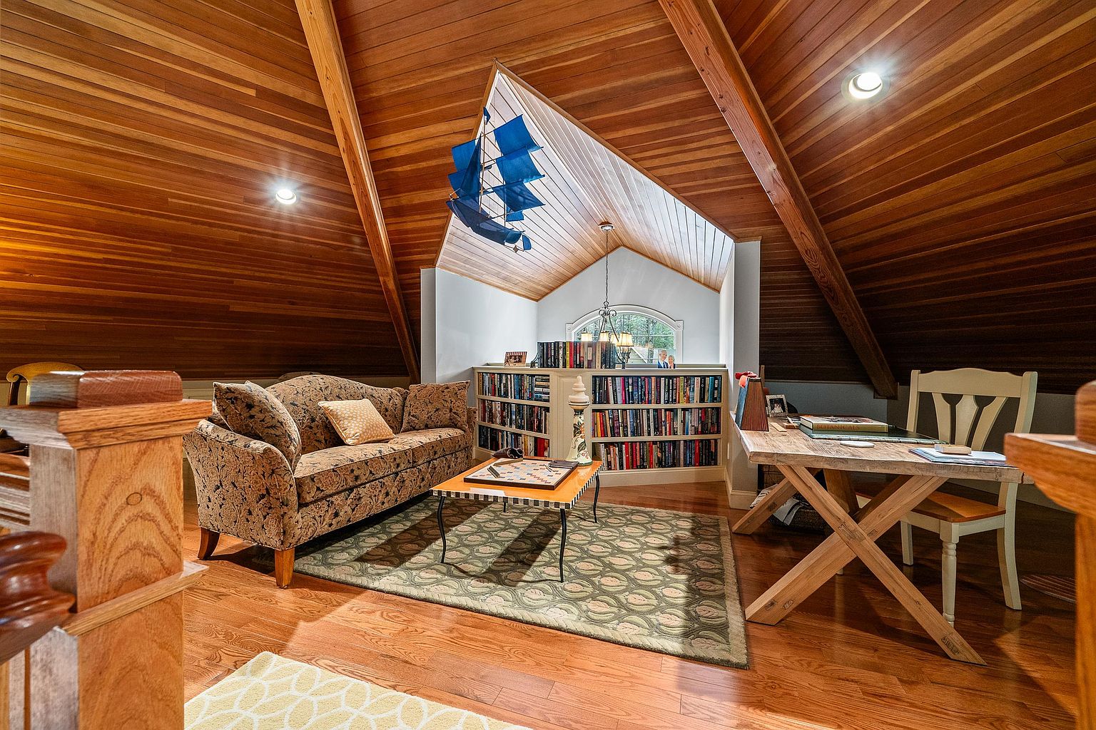 This cozy attic space features warm wood-paneled vaulted ceilings and hardwood floors, creating a rustic and inviting atmosphere. The room is furnished with a patterned sofa, a central coffee table, and a dedicated desk area, all arranged around a built-in bookshelf that serves as a focal point. A decorative model ship hangs from the ceiling, adding a nautical touch to this charming, multi-functional living area.