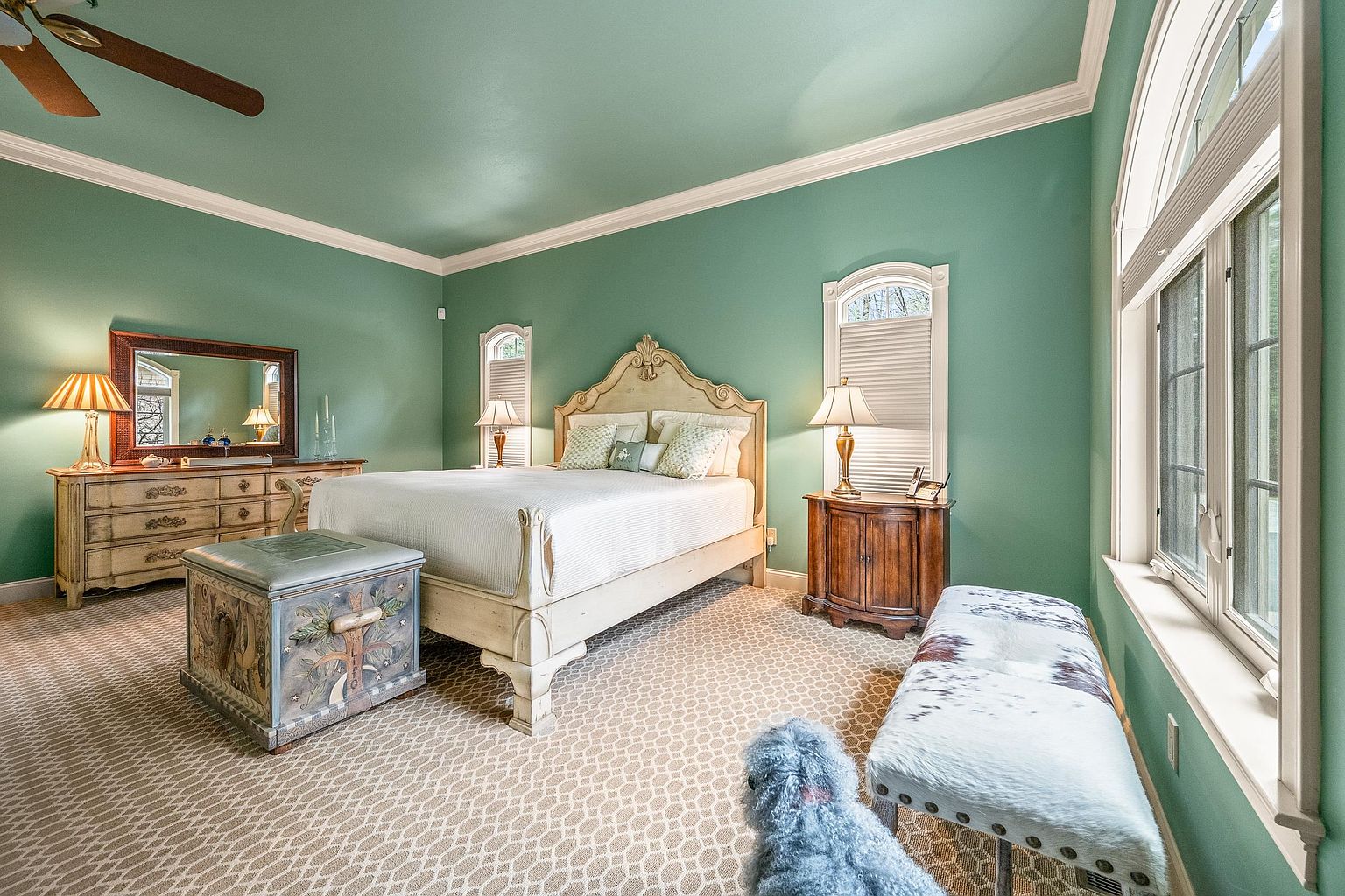 This spacious primary bedroom features soft green walls, a light-colored wooden bed frame, and a patterned carpet, creating a serene and inviting atmosphere. The room is well-lit by two bedside lamps and large windows that allow for plenty of natural light, while a decorative storage chest and a cowhide-patterned bench add unique character. The perspective is a wide-angle shot from the foot of the bed, capturing the entire layout and the elegant, traditional design of the space.