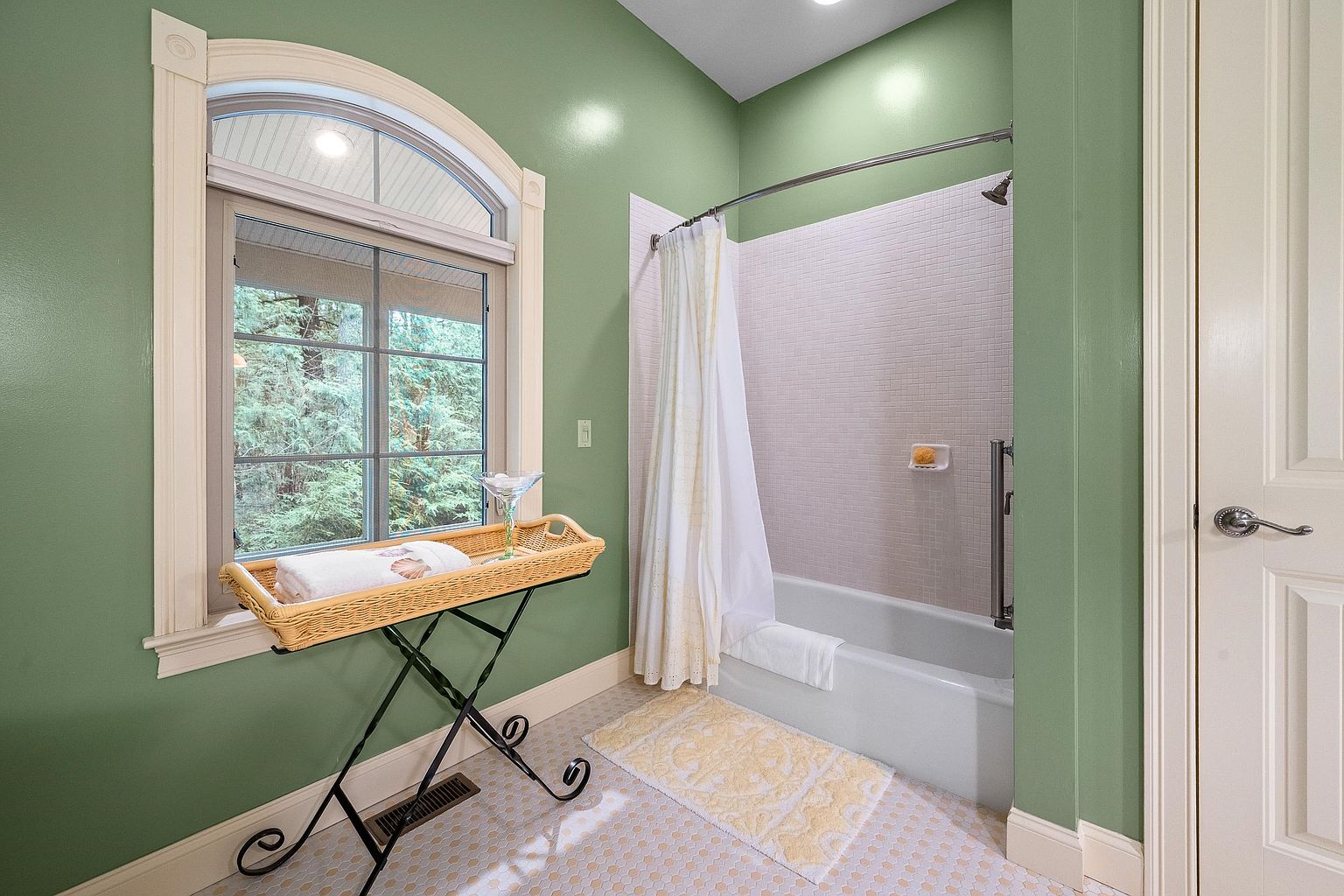 This bathroom features sage green walls, a white tiled shower-tub combination, and a classic arched window that brings in natural light. A decorative wicker tray on a metal stand adds a touch of charm, while the light-colored hexagonal floor tiles create a clean, cohesive look. The space feels bright, airy, and well-maintained, offering a serene atmosphere.