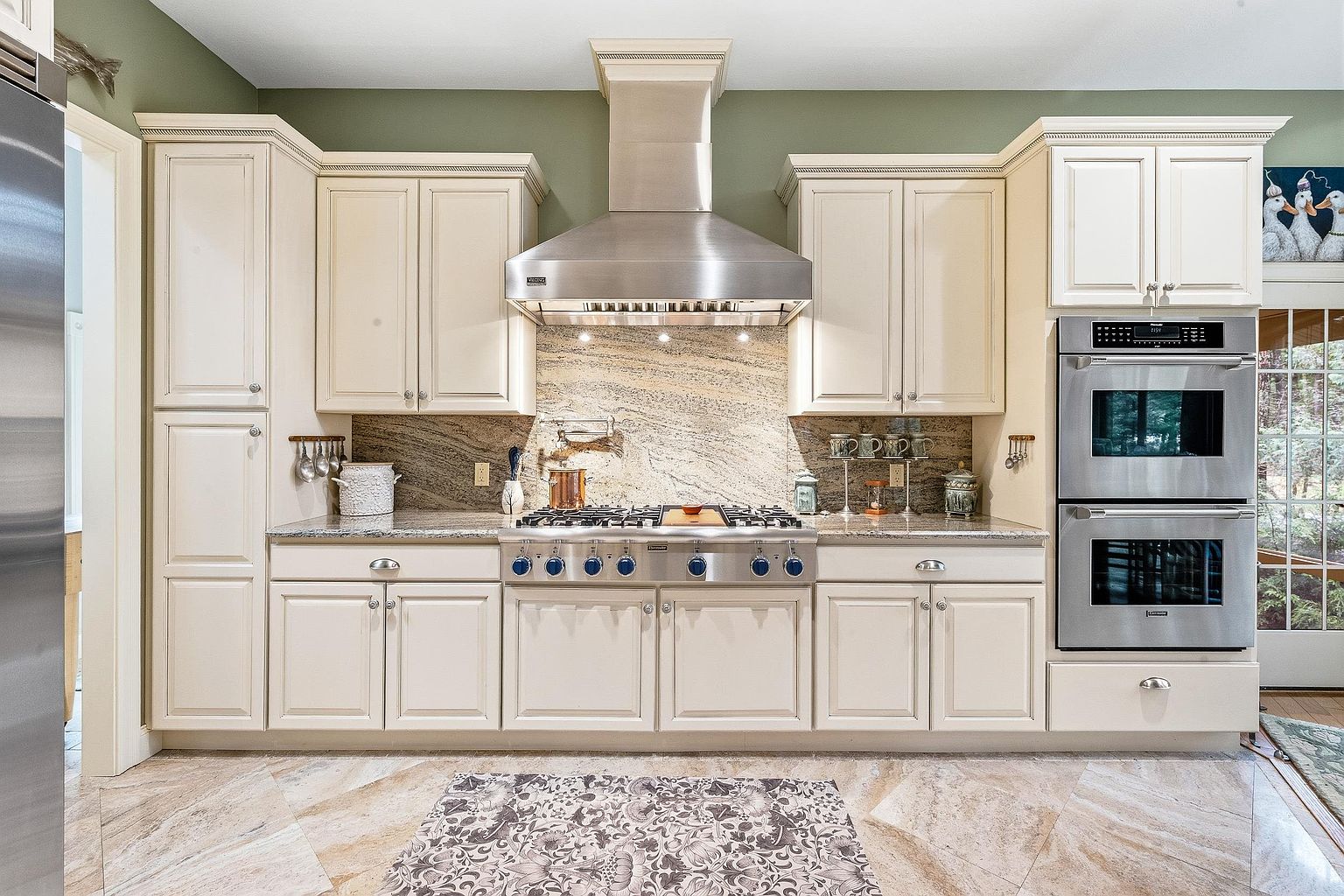 This elegant kitchen features cream-colored cabinetry, a professional-grade stainless steel range with a matching hood, and a stunning granite backsplash. The space is anchored by a double wall oven and polished stone flooring, creating a sophisticated and functional culinary environment. The perspective is a straight-on, eye-level shot that highlights the symmetry and high-end finishes of the kitchen layout.