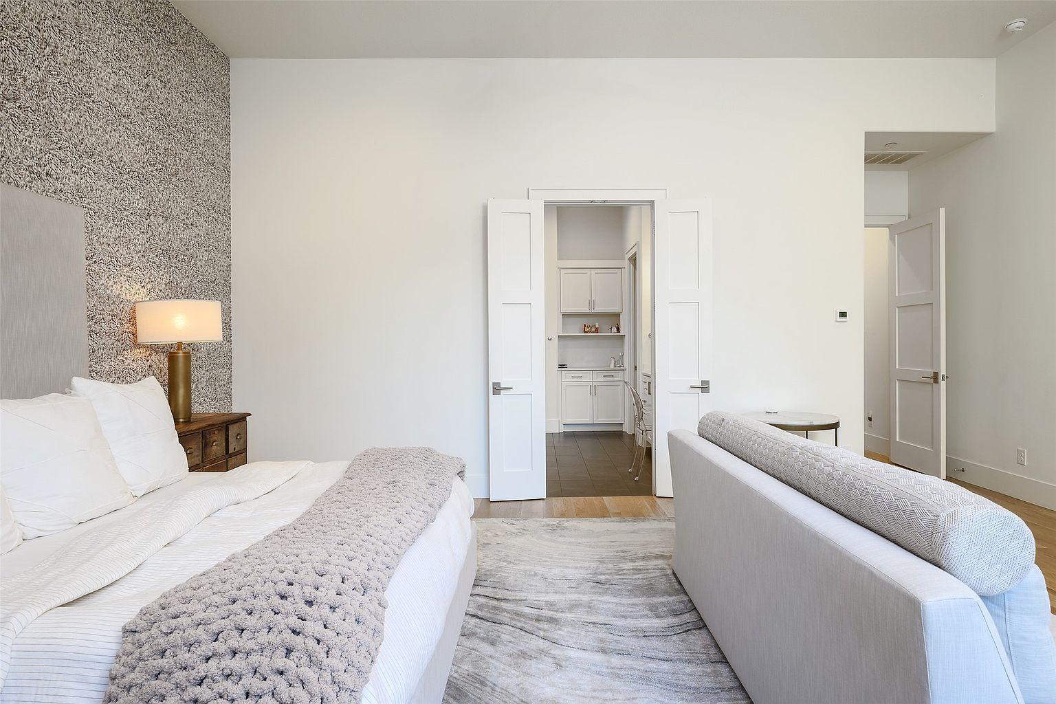 This is a primary bedroom featuring a bed with white linens and a gray throw blanket, a textured accent wall, and a doorway leading to another room. The room has a neutral color palette with white walls and light wood flooring, creating a serene and inviting atmosphere. A gray rug adds texture and ties the space together.
