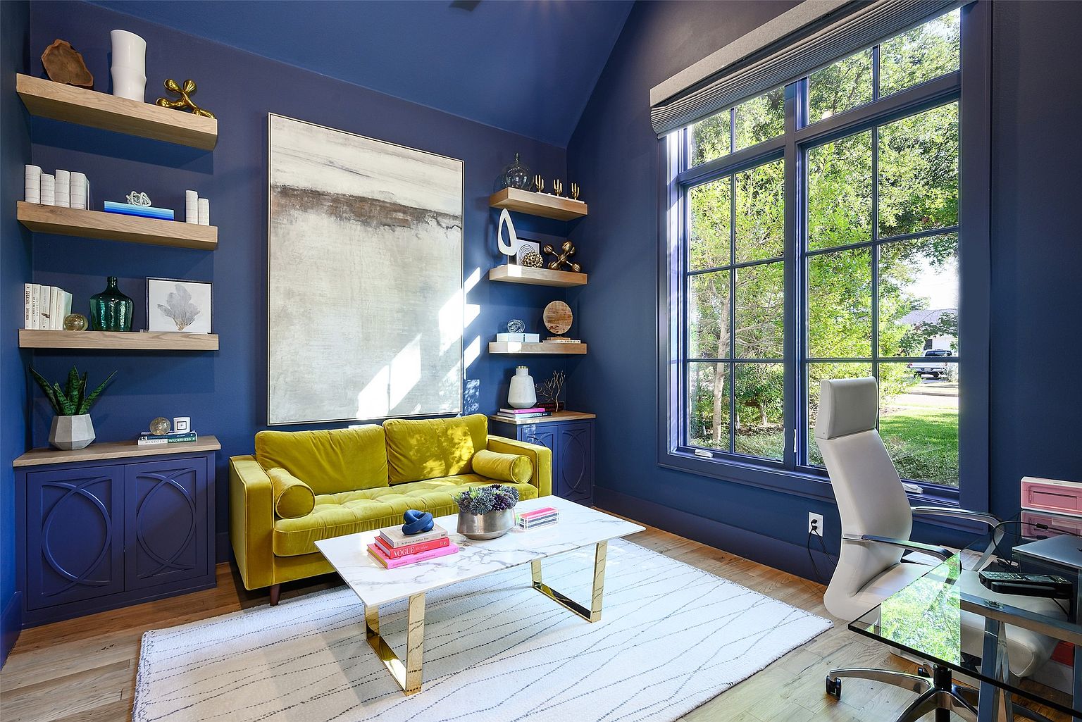 This is an interior shot of a stylish home office or study. The room features a bold blue color scheme, complemented by wooden floating shelves, a large abstract painting, and a vibrant yellow sofa. A large window provides natural light, and the room is furnished with a modern marble coffee table and a glass-topped desk, creating a sophisticated and comfortable workspace.