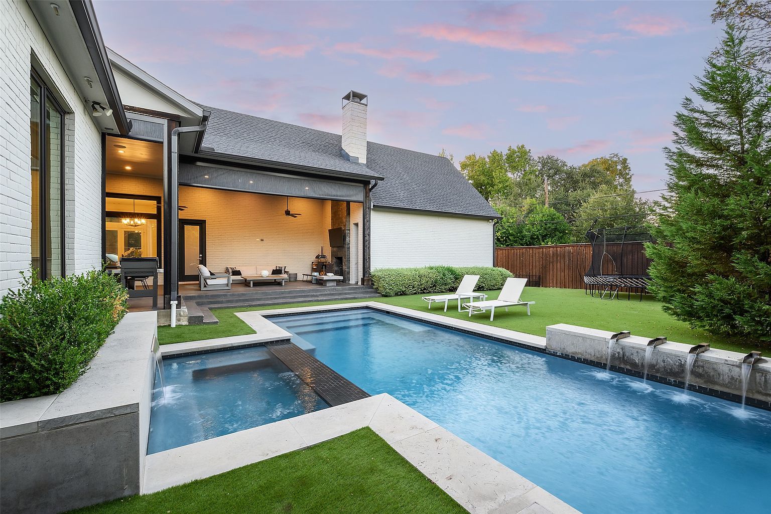This image showcases a luxurious backyard featuring a modern pool and spa area. The pool is surrounded by well-manicured grass and stone accents, with a water feature adding to the ambiance. A covered patio with outdoor seating provides a comfortable space for relaxation and entertainment, enhancing the property's appeal for outdoor living.