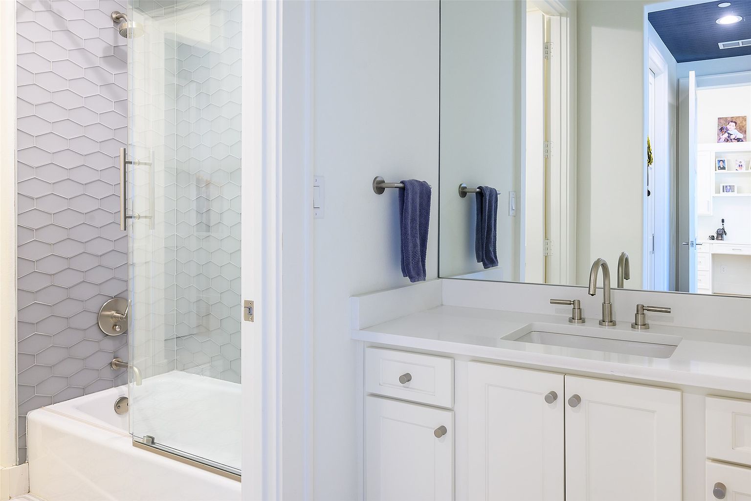 This is a bright and clean bathroom featuring a white vanity with a quartz countertop, modern faucet fixtures, and a large mirror. A shower-tub combination with glass doors and unique gray tiling is visible to the left. The overall impression is fresh and well-maintained, suggesting a comfortable and stylish space.