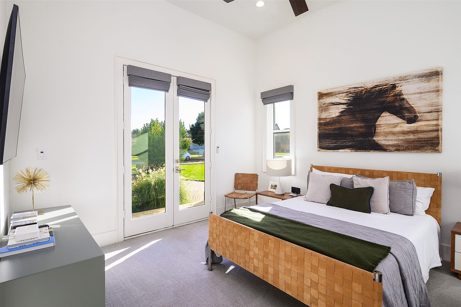 This is a bright and airy bedroom featuring a woven leather bed frame with neutral bedding and decorative pillows. Natural light floods the room through large glass doors and a window, both adorned with gray roman shades. A modern gray console table sits against the wall, complementing the light gray carpet and creating a serene and inviting atmosphere.