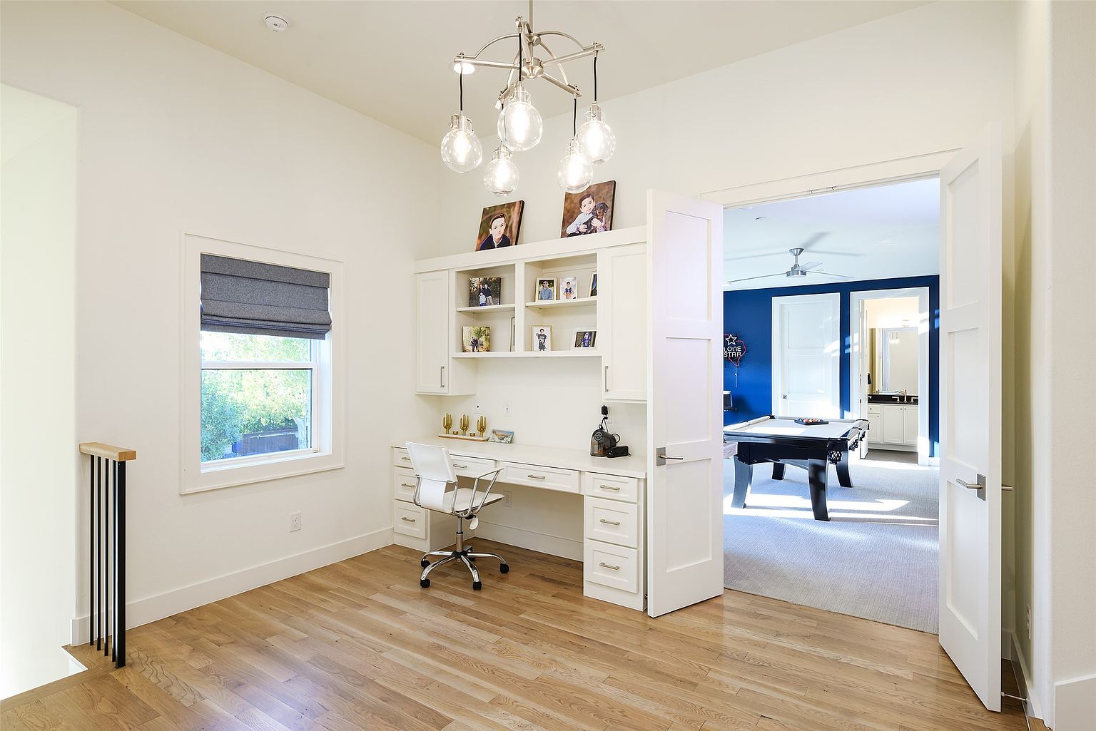 This is an interior shot of a home office featuring a built-in desk with shelving and cabinetry, a window with a gray roman shade, and a modern chandelier. The room has light hardwood floors and white walls, creating a bright and airy atmosphere. An open doorway leads to another room with a pool table, suggesting a recreational space nearby.