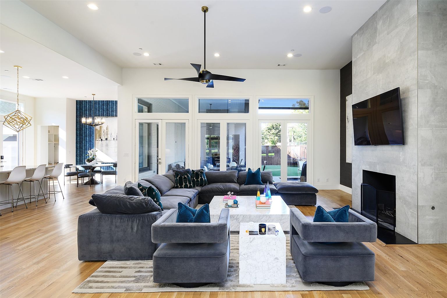 This is a spacious and modern living room featuring a large sectional sofa, two armchairs, and a marble coffee table. The room has hardwood floors, a fireplace with a gray stone surround, and large windows that provide ample natural light. The overall style is contemporary and luxurious, creating a comfortable and inviting space.