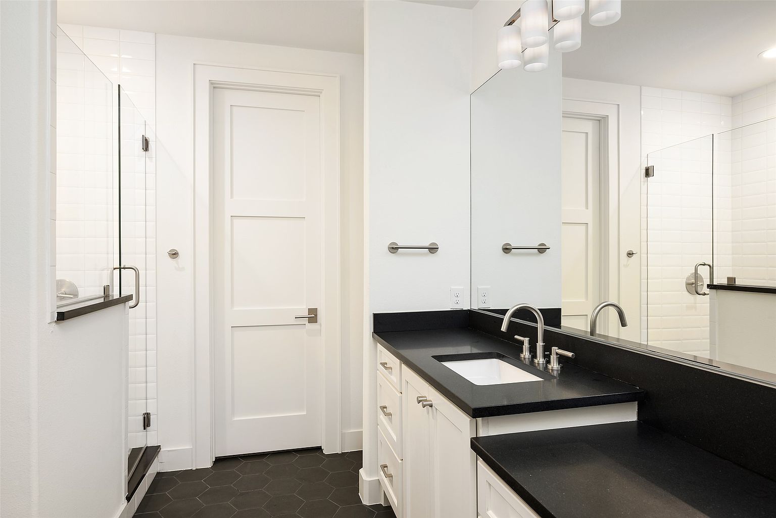 This is a well-lit bathroom featuring a modern design with a black countertop and white cabinetry. The floor is covered in dark, hexagonal tiles, and a glass-enclosed shower is visible to the left. A large mirror reflects the space, enhancing the sense of openness and cleanliness.
