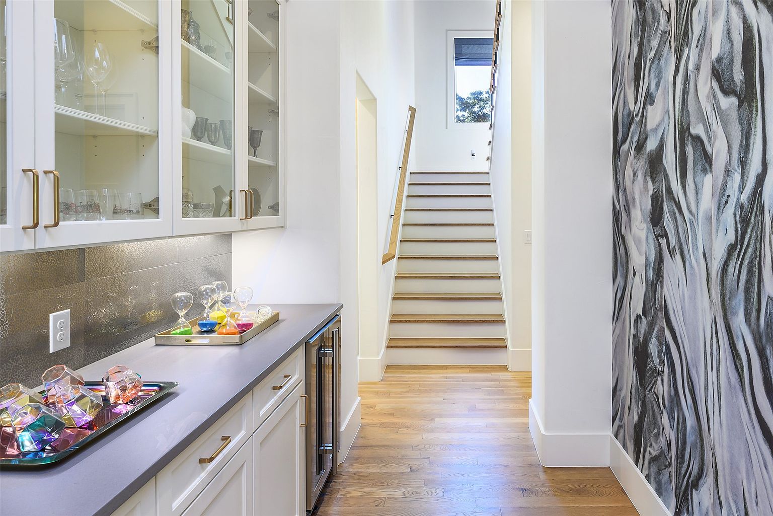 This interior shot showcases a hallway with a stylish bar area on the left and a staircase leading upwards. The bar features white cabinetry with glass doors, a gray countertop, and a metallic backsplash, while the hallway is adorned with a striking black and white patterned wallpaper on the right. The hardwood flooring and white walls create a bright and modern aesthetic.