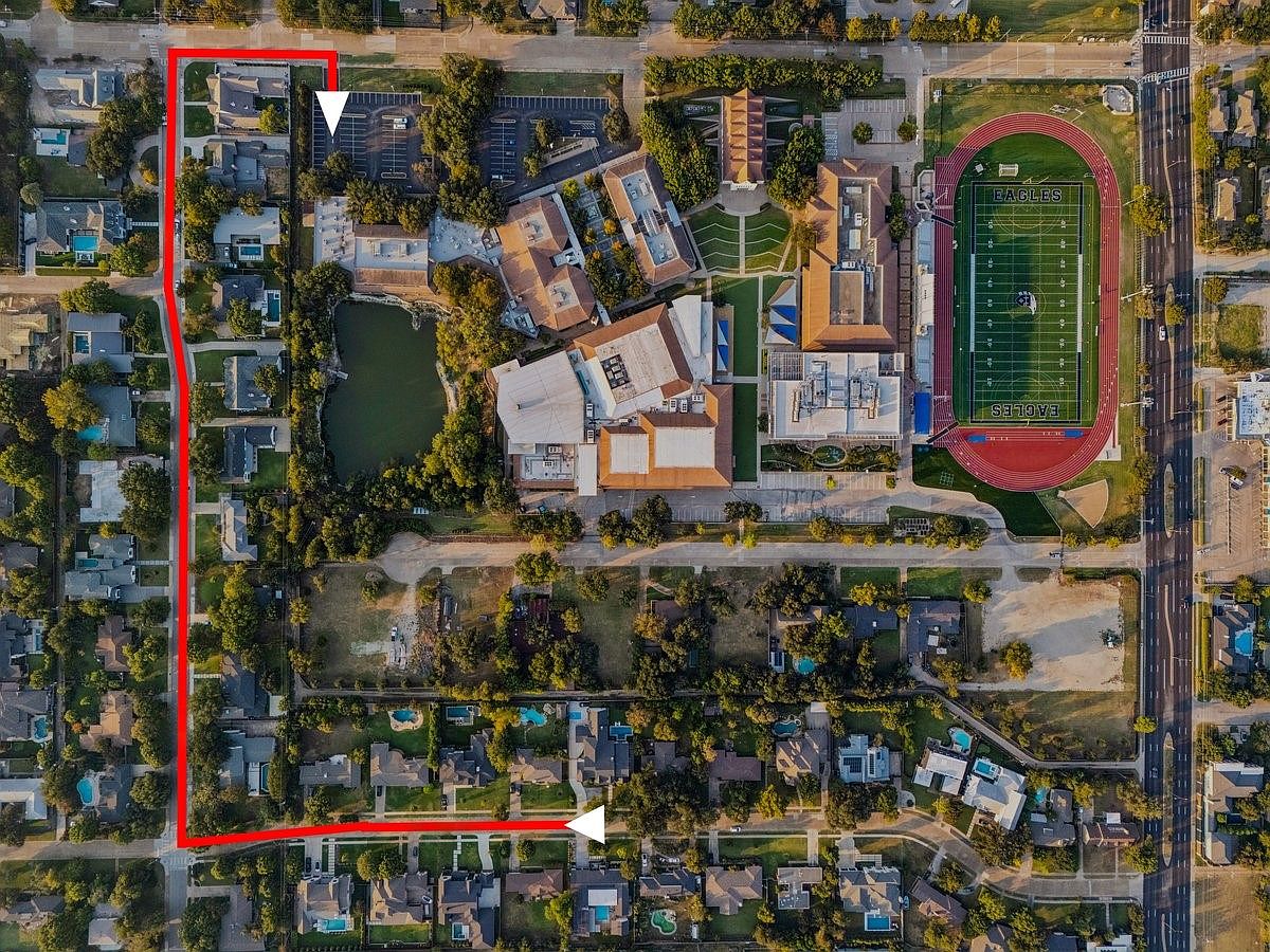 This aerial view showcases a residential area with a mix of houses, a school complex featuring a football field, and a small lake. The properties are surrounded by mature trees, and the layout suggests a well-established neighborhood. A red line highlights a specific area of interest, possibly indicating property boundaries or a route.