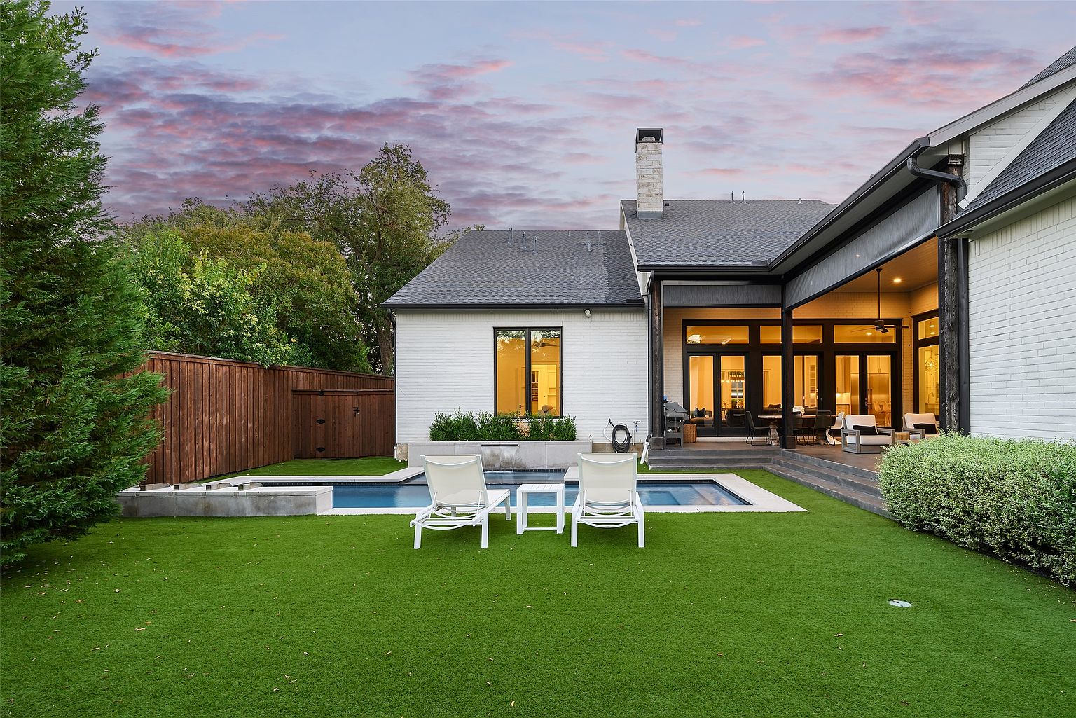 This image showcases the backyard of a modern home, featuring a rectangular pool with a minimalist design, surrounded by artificial turf. Two white lounge chairs and a small table are positioned poolside, inviting relaxation. The house, painted in white brick, has a covered patio area with outdoor furniture, and the overall impression is one of luxury and tranquility.