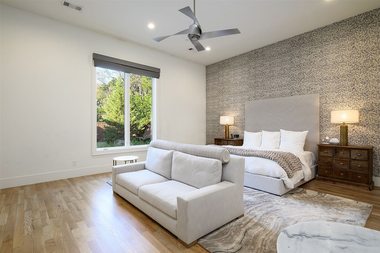 This is a well-lit primary bedroom featuring a large bed with a neutral-toned headboard and crisp white linens. The room includes a comfortable sofa, wooden nightstands with lamps, and a textured accent wall behind the bed. The hardwood floors and area rug add warmth and style to the space, creating a serene and inviting atmosphere.