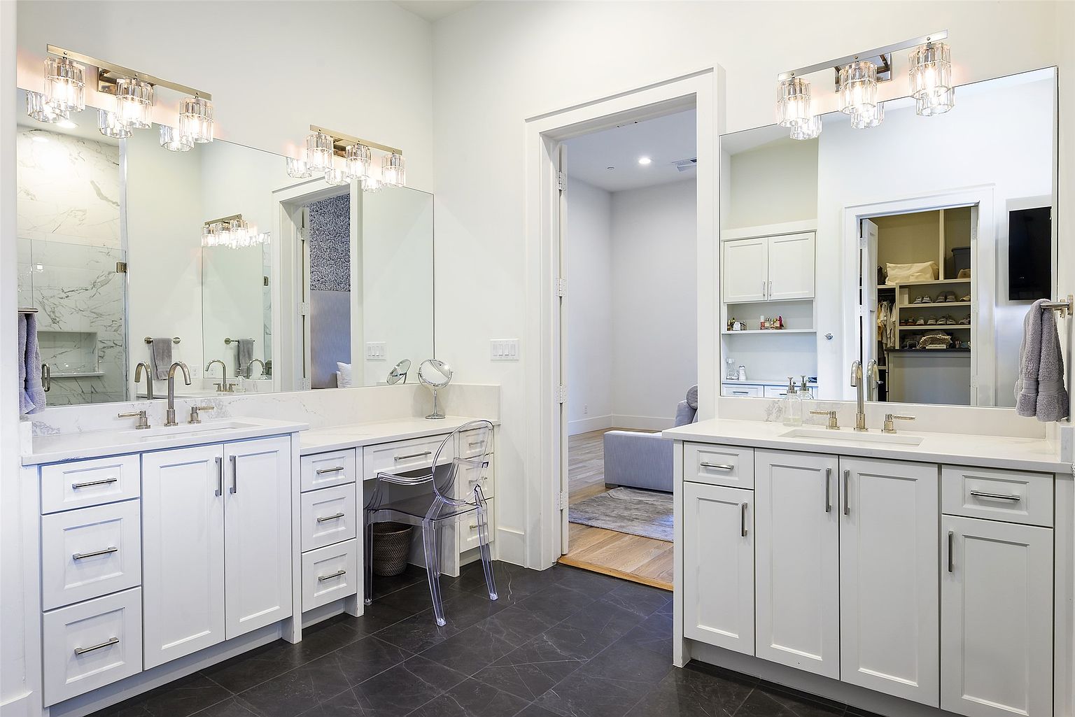 This is a well-lit primary bathroom featuring dual vanities with white cabinetry and countertops, large mirrors, and modern light fixtures. The dark tile flooring contrasts with the bright white walls and cabinets, creating a clean and sophisticated look. A clear acrylic chair sits at the vanity, and an open doorway leads to another room, possibly a dressing area or closet.