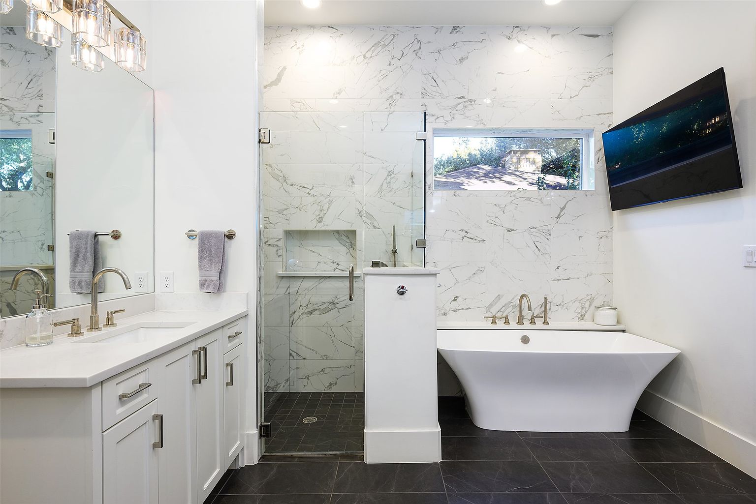 This is a primary bathroom featuring a modern design with white marble tile walls and dark gray tile flooring. The bathroom includes a freestanding white bathtub, a glass-enclosed shower, and a vanity with white cabinets and a light countertop. A wall-mounted television adds a touch of luxury.