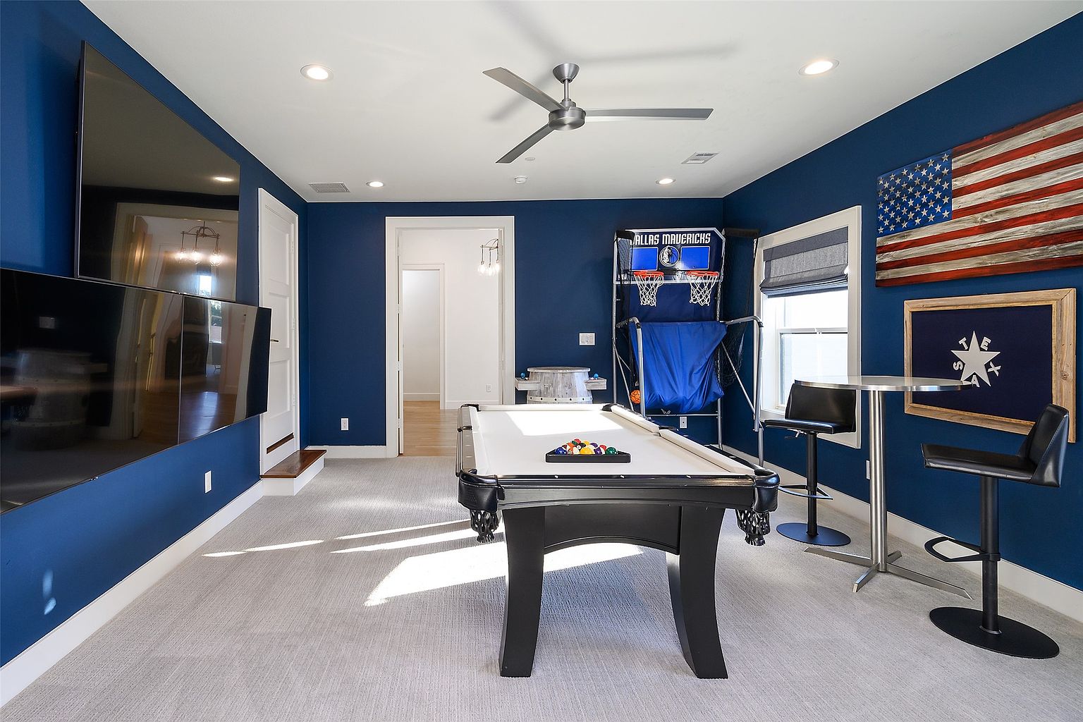 This is a basement game room featuring a pool table, a basketball arcade game, and a bar area with stools. The walls are painted a deep blue, and there's an American flag and a Texas flag hanging on the wall. The room is well-lit with recessed lighting and a ceiling fan, creating a fun and inviting atmosphere.