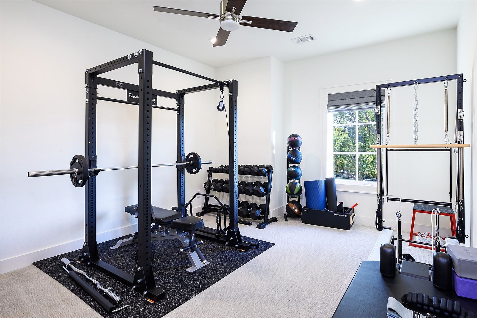 This is a well-equipped home gym featuring a power rack with a barbell and weight plates, an adjustable bench, and a dumbbell rack. A pilates reformer is positioned near a window, and medicine balls are neatly stacked. The room has a clean and modern aesthetic with white walls and a neutral-colored carpet.