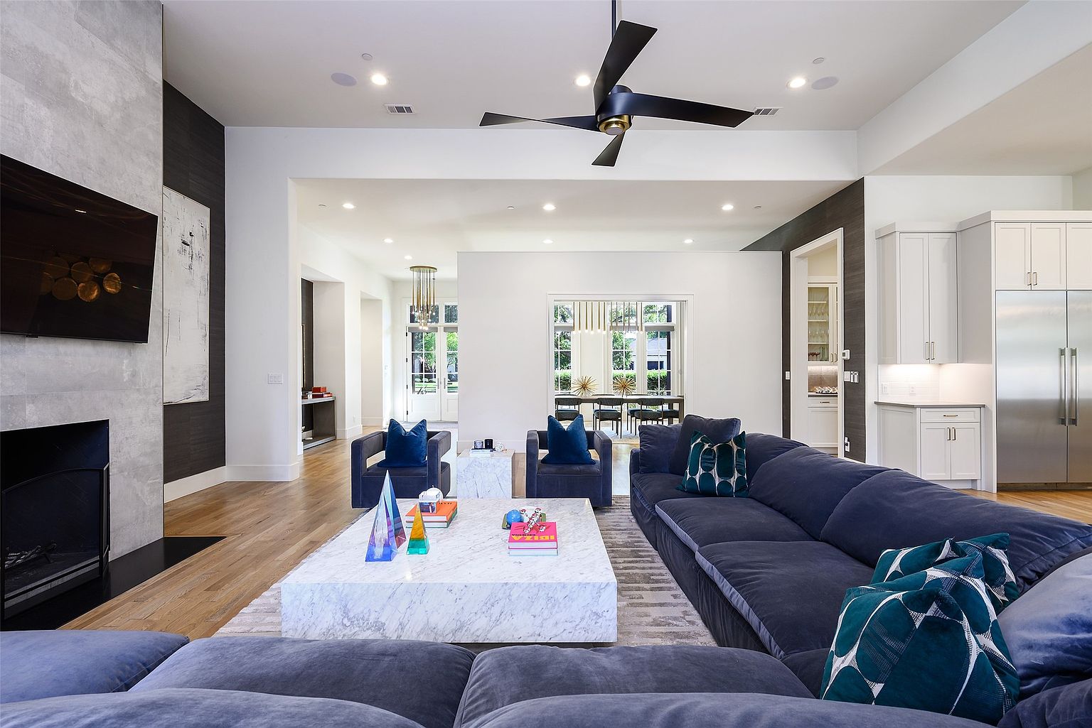 This is a spacious and luxurious living room featuring a large, plush blue sectional sofa and two armchairs. A marble coffee table sits in the center of the room, adding a touch of elegance. The room is well-lit with recessed lighting and natural light, creating a bright and inviting atmosphere, and the open floor plan seamlessly connects to other areas of the home.