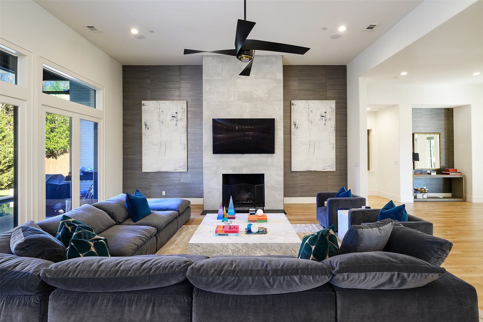 This is a spacious living room featuring a large, plush sectional sofa in a dark gray color, complemented by blue and patterned throw pillows. A modern fireplace with a mounted television is the focal point, flanked by abstract artwork. The room is well-lit with recessed lighting and natural light from a large window, showcasing a contemporary and comfortable living space.
